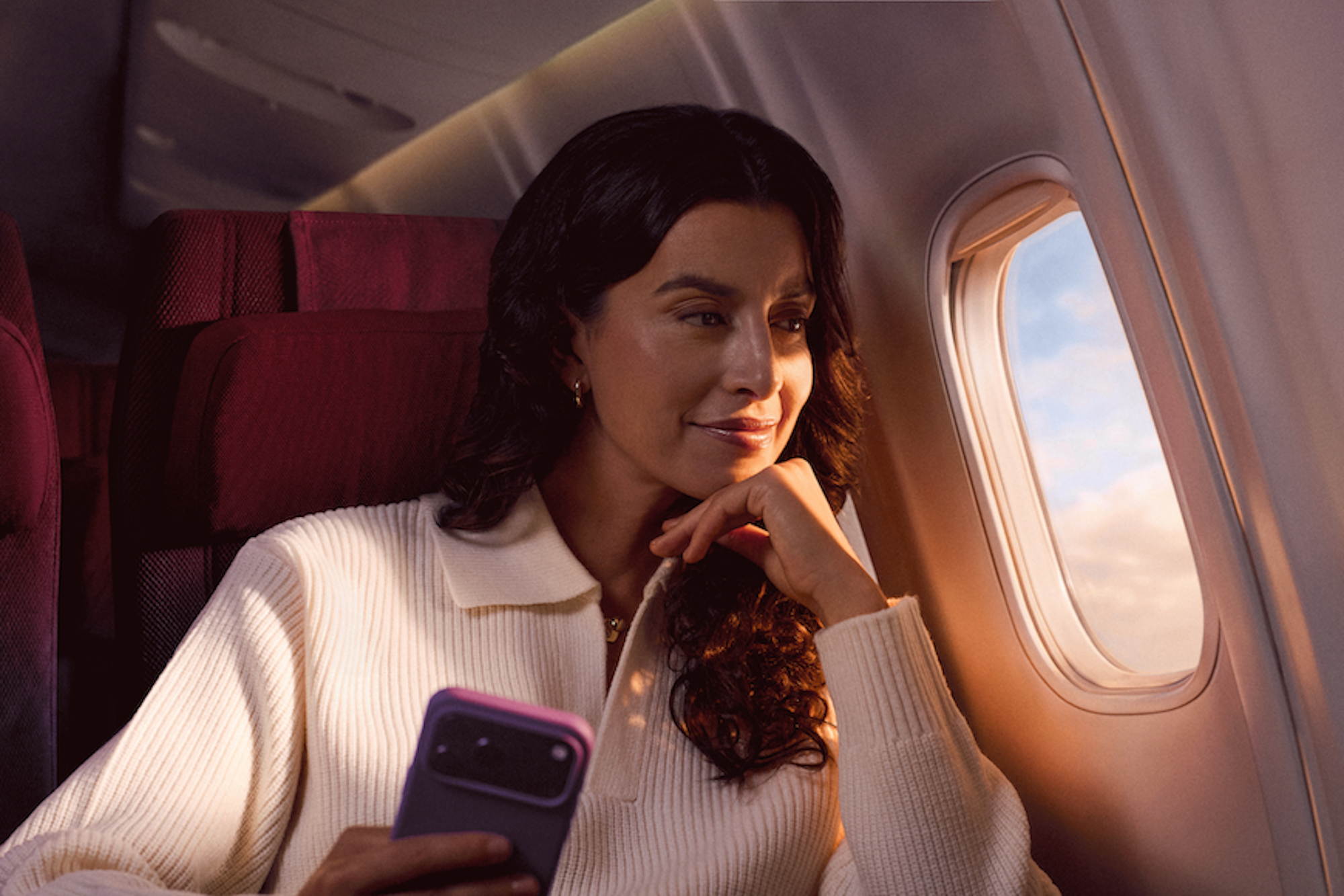 Mulher segurando um celular com capinha roxa. Ela está no avião, sentada próxima à janela e está usando uma blusa de tricô branca.