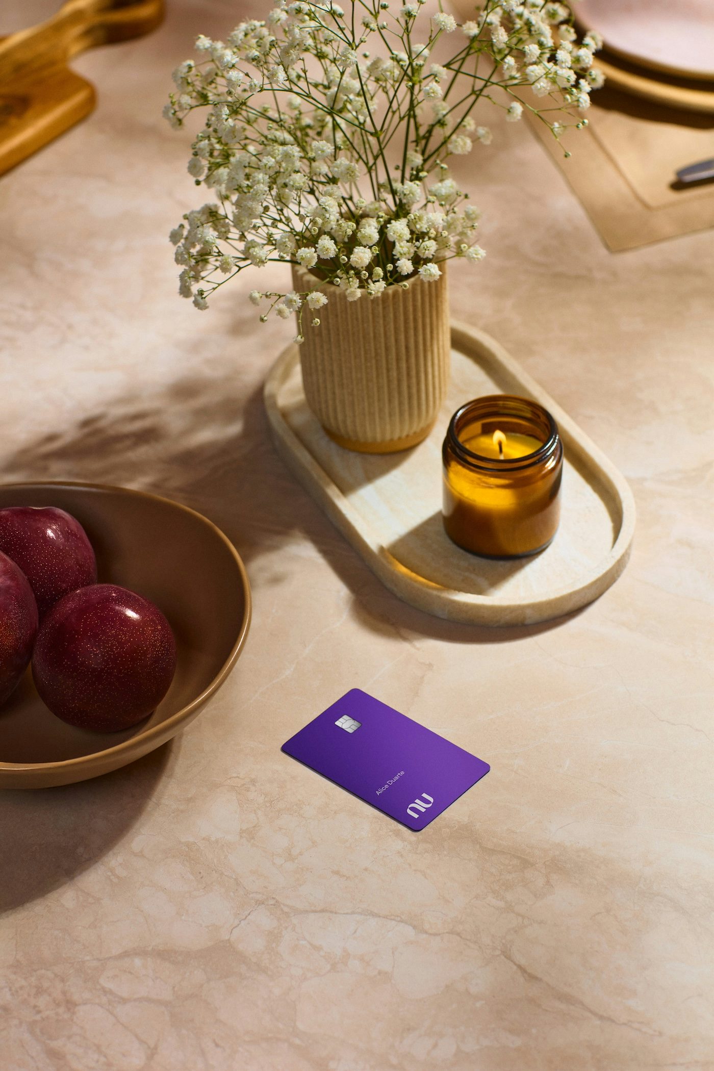 O cartão Nubank Ultravioleta está posicionado em primeiro plano, acompanhado por uma tigela com ameixas vermelhas, uma vela aromática acesa e um vaso com delicadas flores mosquitinho.