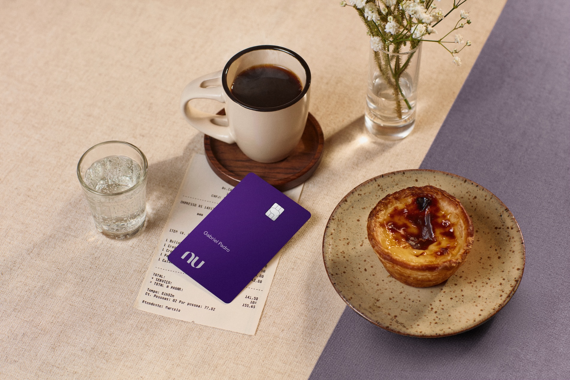 Mesa posta com um pastel de nata em um prato de cerâmica, uma caneca de café preto sobre pires de madeira, um copo de água e um pequeno vaso com flores brancas. O cartão Nubank Ultravioleta aparece em destaque sobre a conta do estabelecimento.