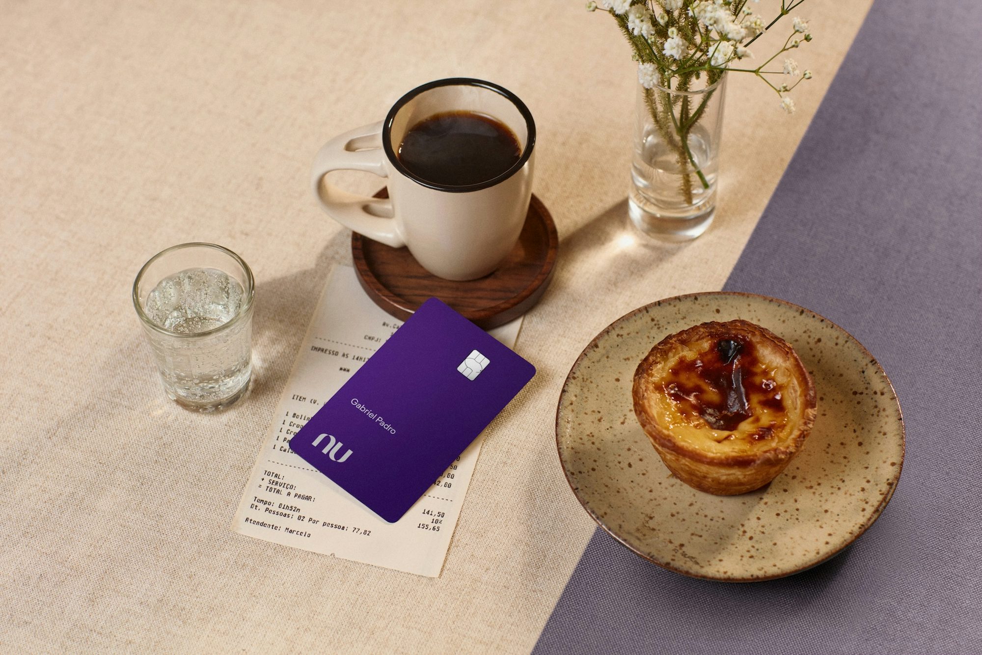 Mesa posta com um pastel de nata em um prato de cerâmica, uma caneca de café preto sobre pires de madeira, um copo de água e um pequeno vaso com flores brancas. O cartão Nubank Ultravioleta aparece em destaque sobre a conta do estabelecimento.