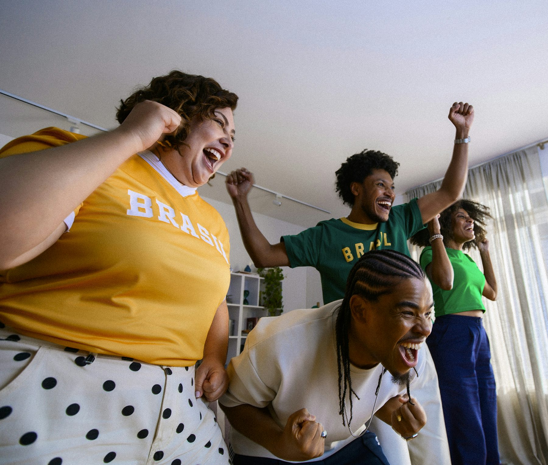 Quatro pessoas comemoram animadamente em uma sala de estar. À esquerda, uma mulher branca de cabelo cacheado usa camiseta amarela com "Brasil" e calça branca de poás. Ao centro, um homem negro com tranças se inclina para frente celebrando. Ao fundo, um homem negro com camiseta verde "Brasil" ergue o punho, e uma mulher negra de top verde sorri. Estante branca e cortinas ao fundo.