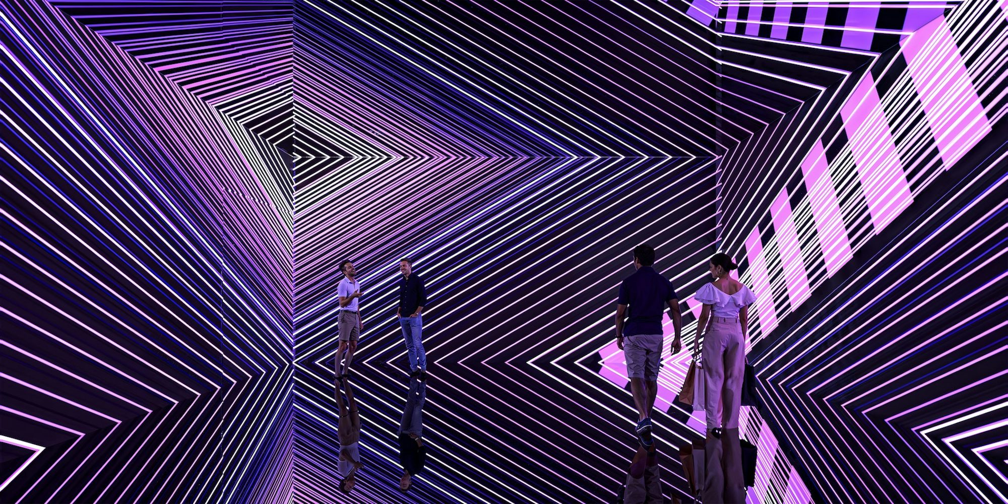 Visitantes em um espaço espelhado preenchido por projeções de linhas geométricas luminosas em tons de roxo, rosa e branco, que geram um forte efeito de ilusão de ótica e profundidade infinita.