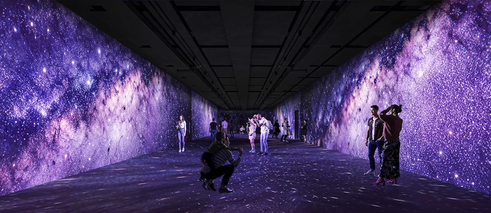 Pessoas caminham e interagem dentro de um longo corredor imersivo com projeções digitais de alta definição que simulam uma galáxia estrelada nas paredes, teto e chão, criando uma atmosfera cósmica e em tons de roxo.