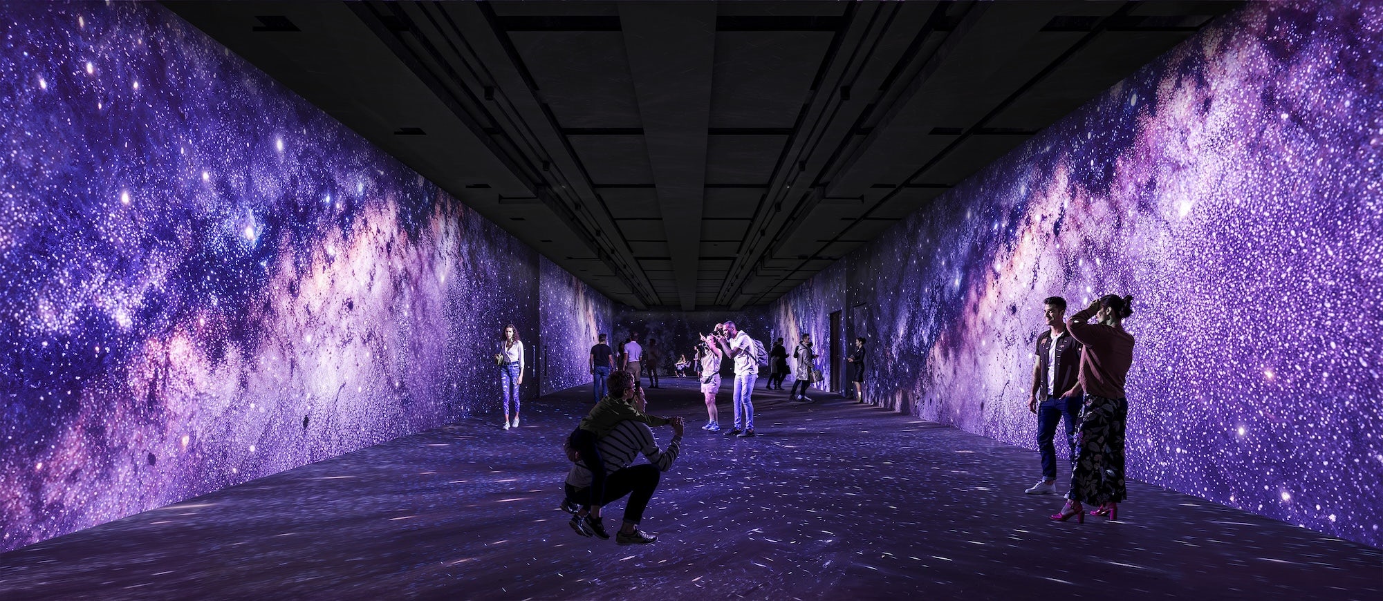Pessoas caminham e interagem dentro de um longo corredor imersivo com projeções digitais de alta definição que simulam uma galáxia estrelada nas paredes, teto e chão, criando uma atmosfera cósmica e em tons de roxo.