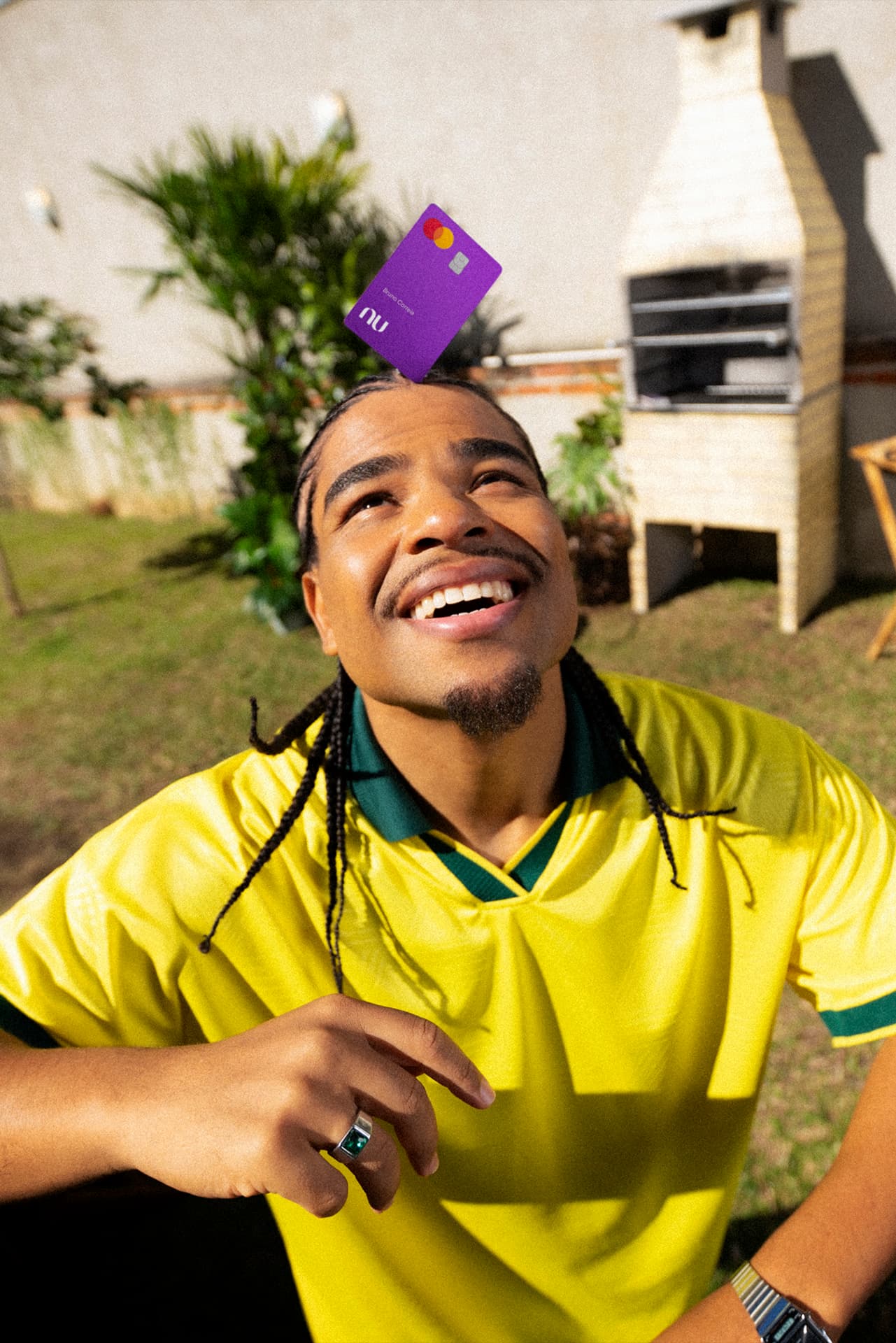 Jovem rapaz equilibra o cartão do Nubank na cabeça como se fizesse embaixadinhas, vestindo uma camiseta verde e amarela em um quintal gramado com churrasqueira ao fundo.