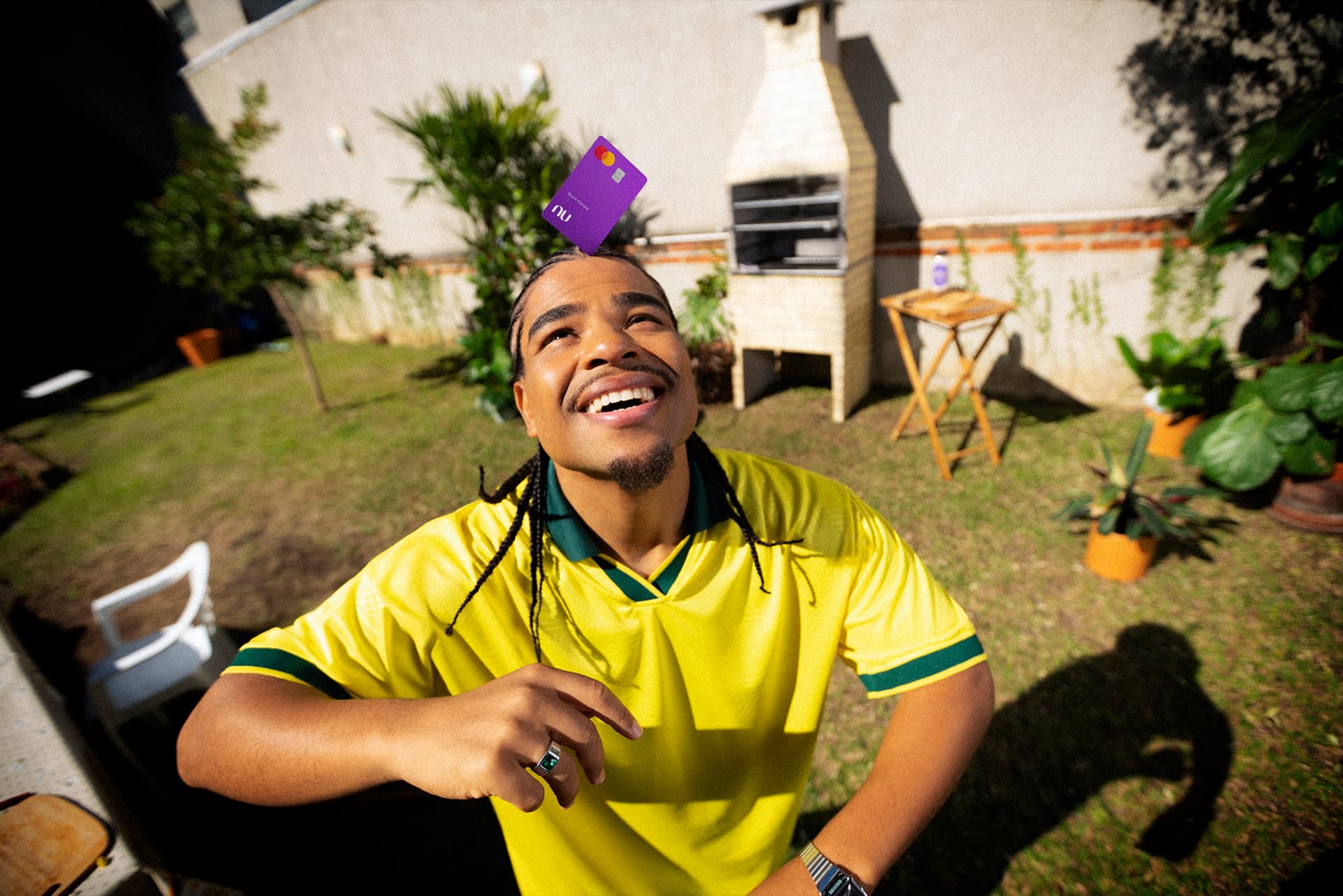 Jovem rapaz equilibra o cartão do Nubank na cabeça como se fizesse embaixadinhas, vestindo uma camiseta verde e amarela em um quintal gramado com churrasqueira ao fundo