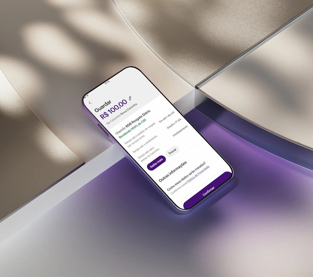 Celular apoiado em uma forma geométrica branca com aplicativo Nubank aberto na área de Caixinhas, guardando 100 reais em uma nova caixinha.