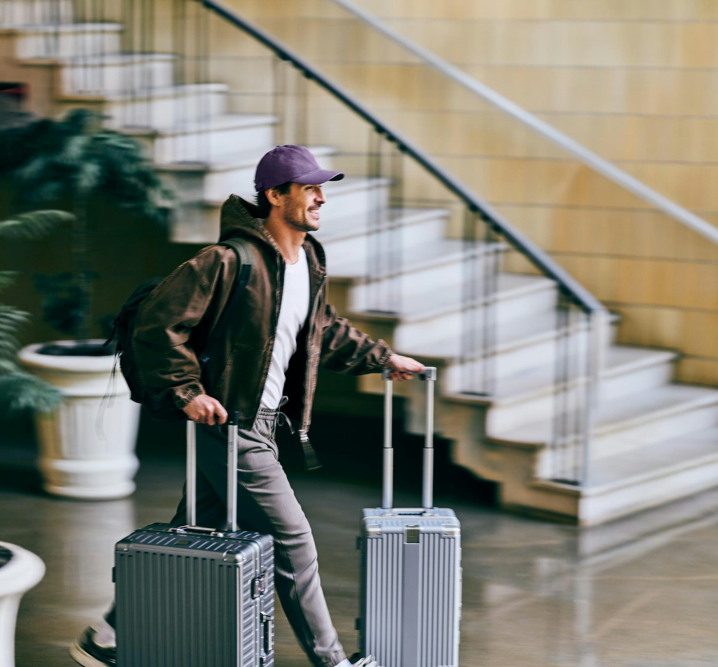 Um homem usando um boné roxo e jaqueta marrom caminha apressado carregando uma mochila nas costas e puxando duas malas de rodinha prateadas.