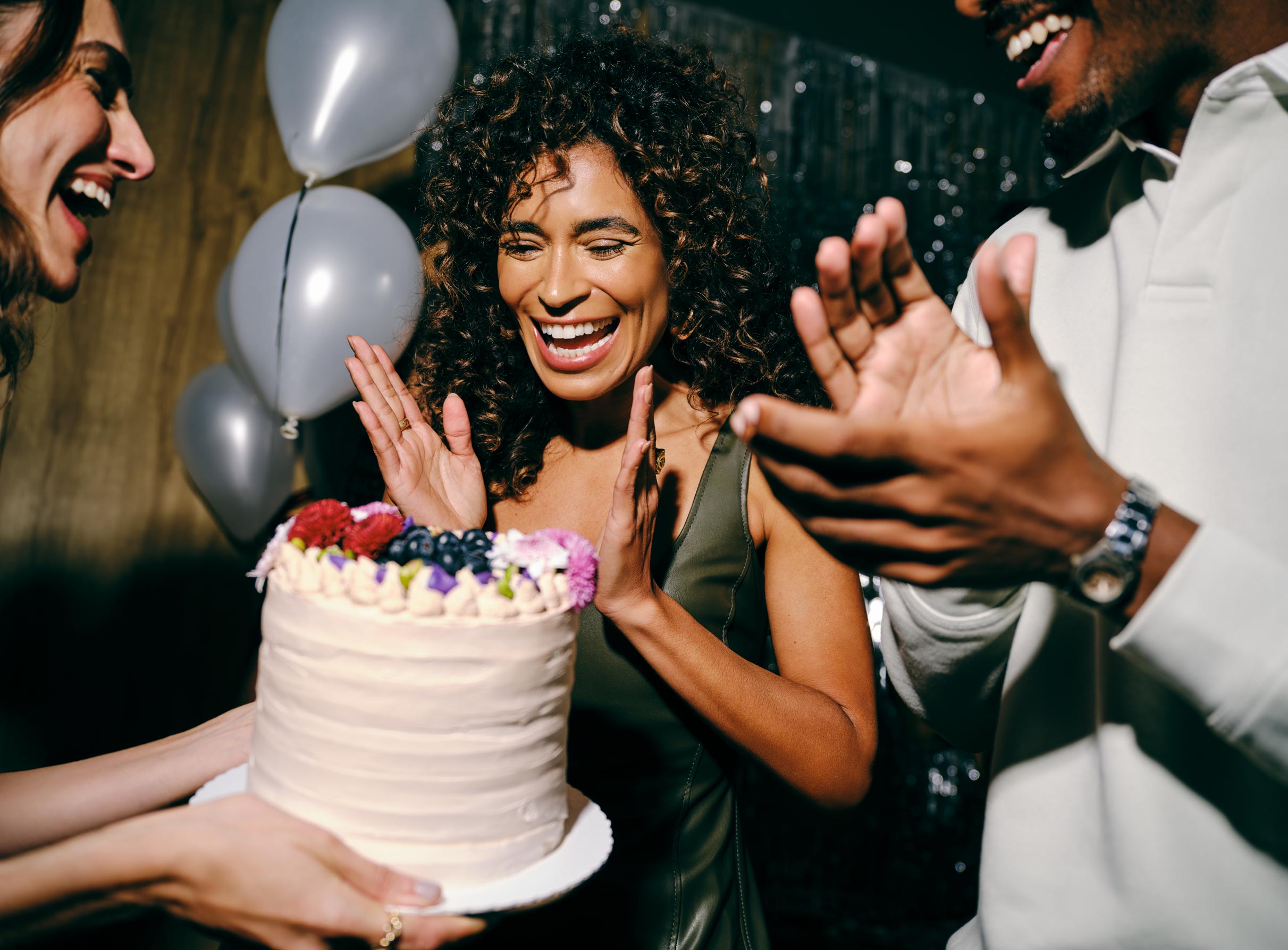 Uma mulher de cabelos cacheados sorri enquanto bate palmas diante de um bolo de aniversário confeitado, com balões prateados decorando o fundo da festa.