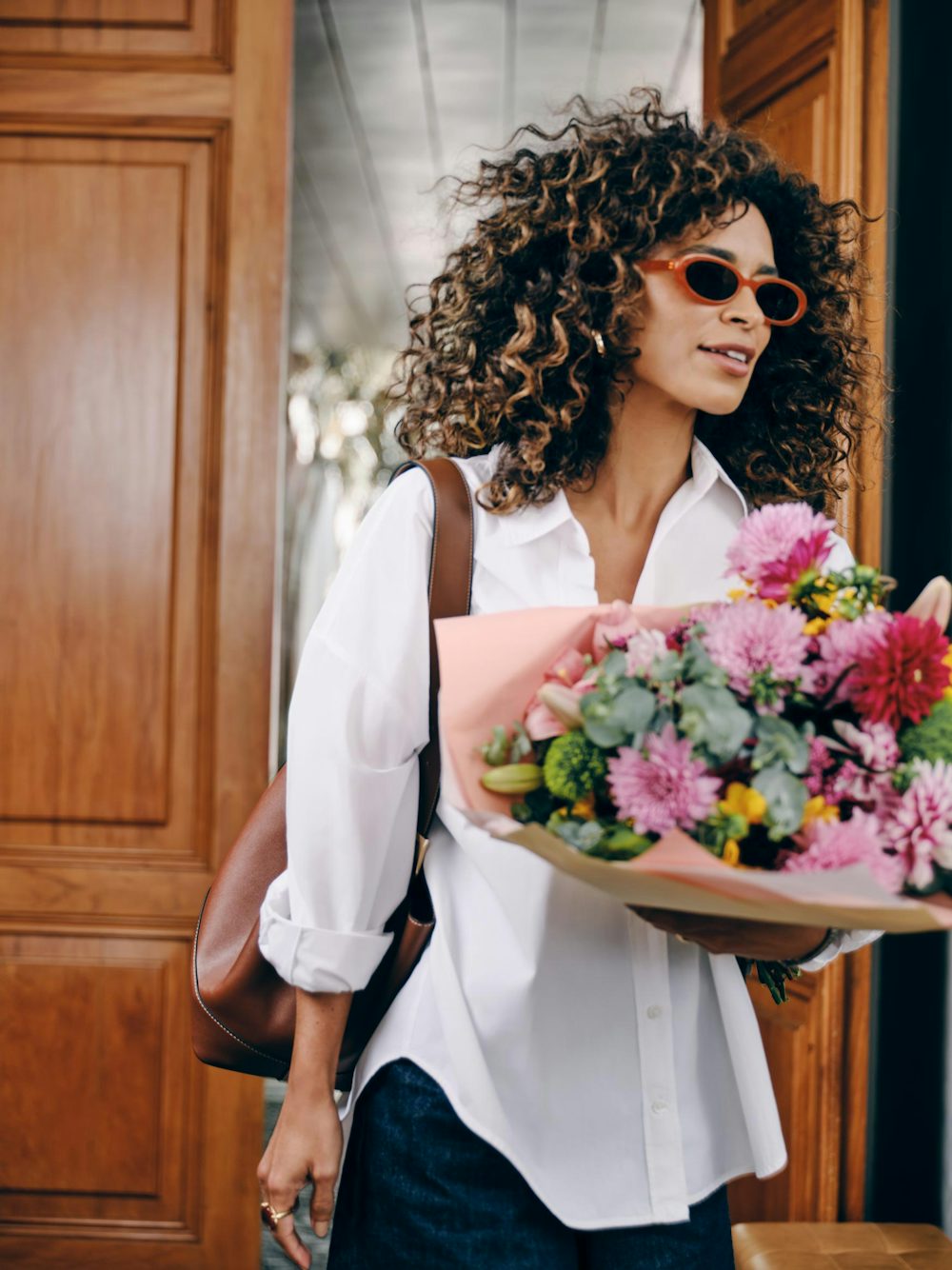 Mulher com cabelo cacheado e óculos, segurando buquê de flores.