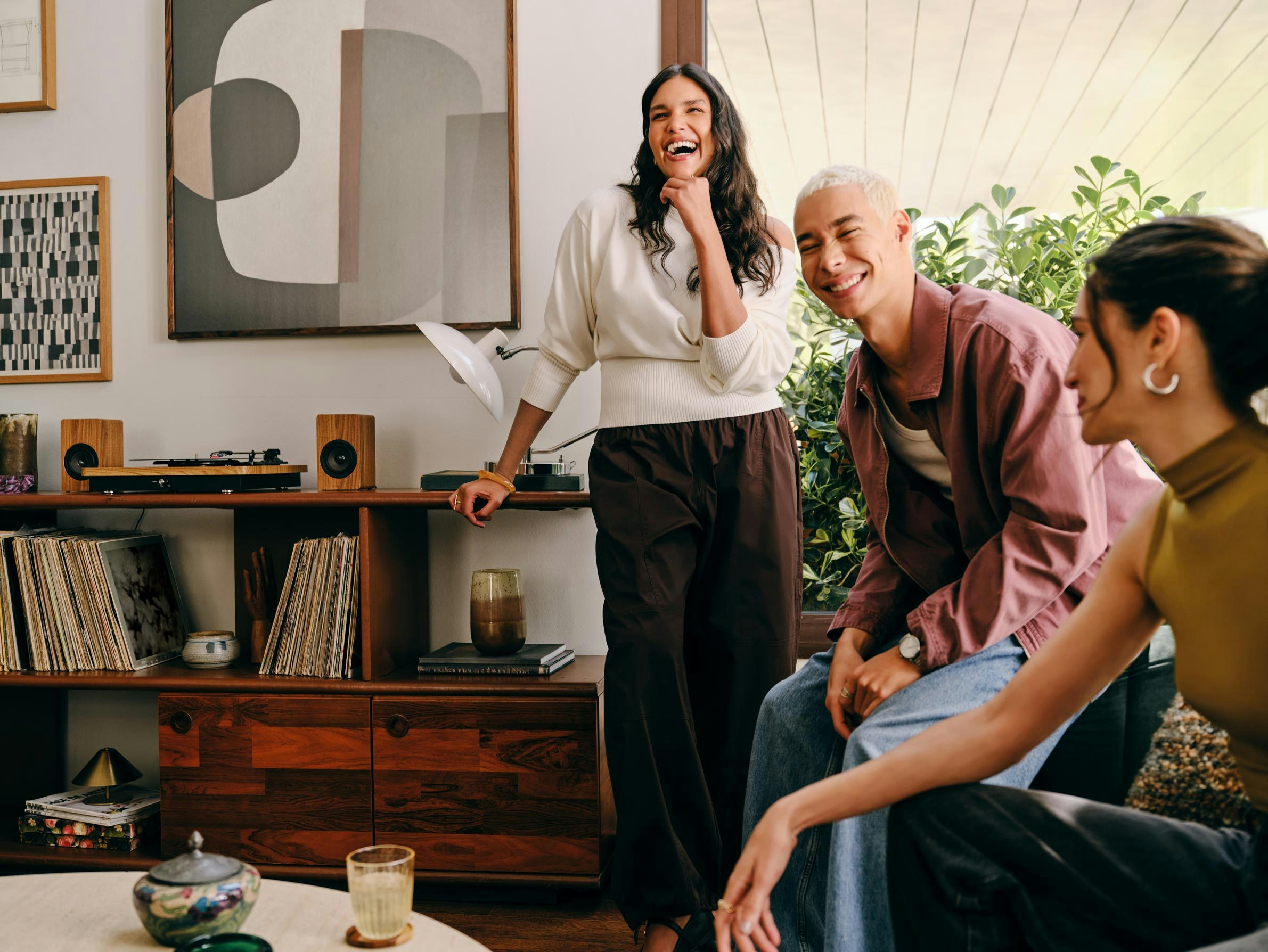 Três amigos interagem de forma descontraída em uma sala de estar. Ao fundo, aparece um móvel de madeira com um toca-discos e vários discos de vinil.