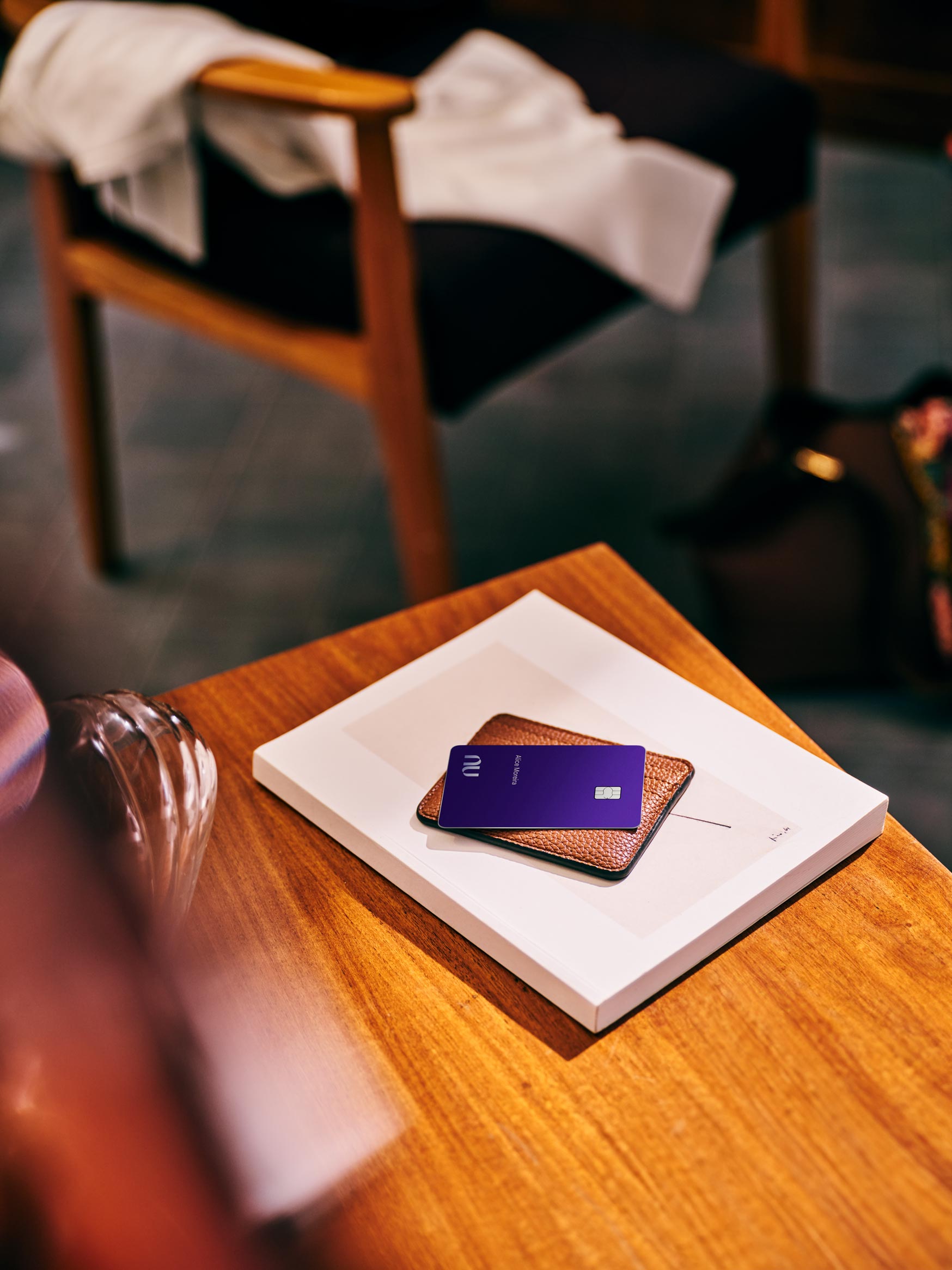 O cartão Nubank Ultravioleta aparece em cima de uma carteira de couro marrom apoiada sobre um livro branco em uma mesa de madeira clara. Uma poltrona com uma manta branca aparece no fundo desfocado.