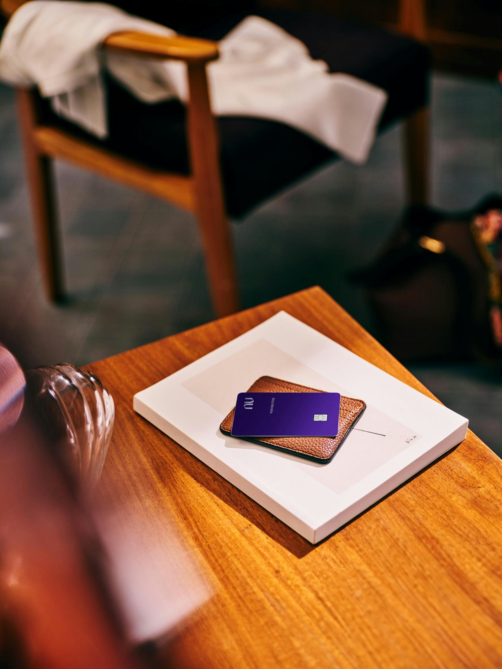 O cartão Nubank Ultravioleta aparece em cima de uma carteira de couro marrom apoiada sobre um livro branco em uma mesa de madeira clara. Uma poltrona com uma manta branca aparece no fundo desfocado.