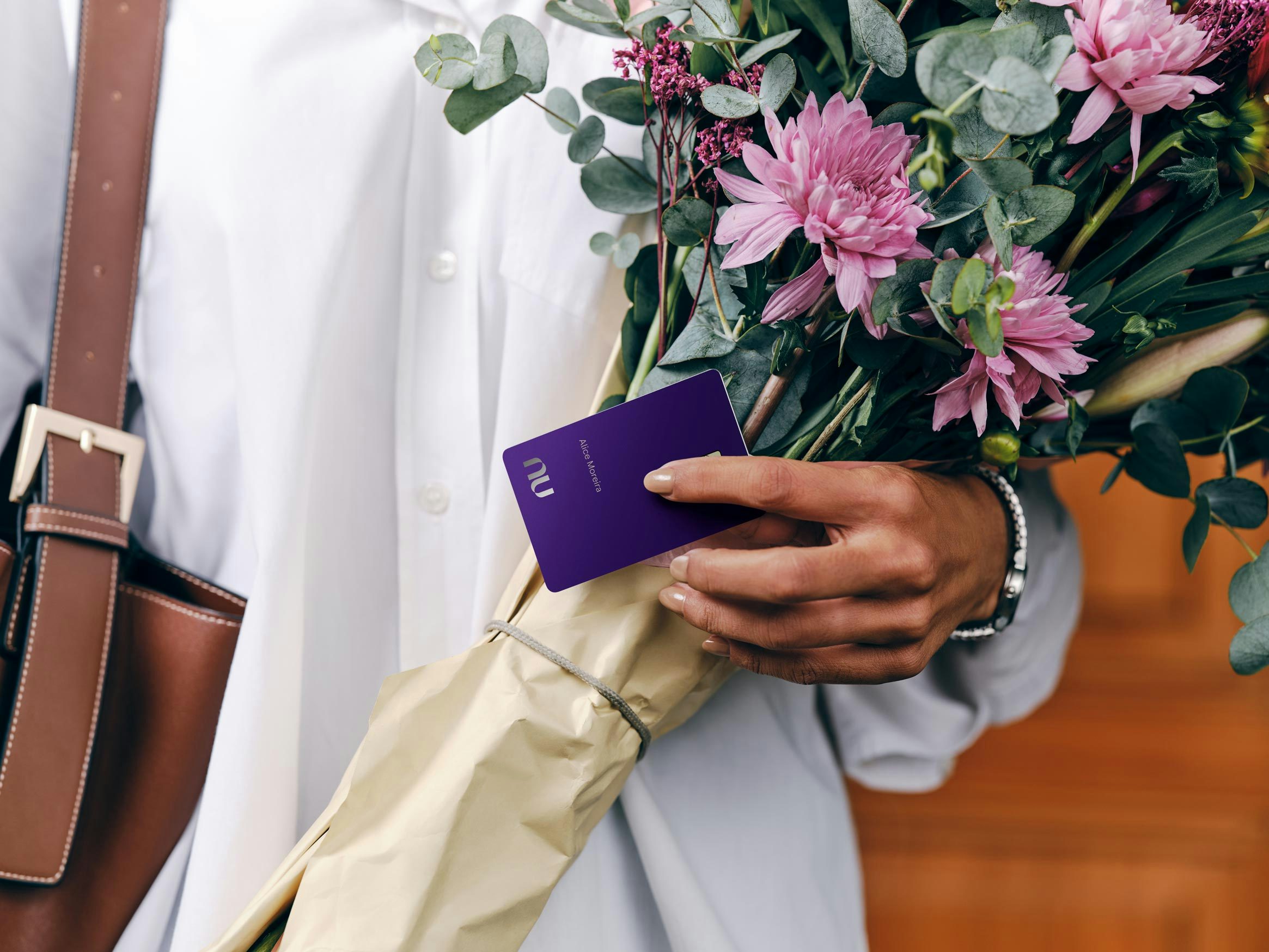 Uma mulher vestindo camisa branca e bolsa de couro marrom, segurando um grande buquê de flores rosas e folhagens. Com a mesma mão, ela exibe o cartão Nubank Ultravioleta.
