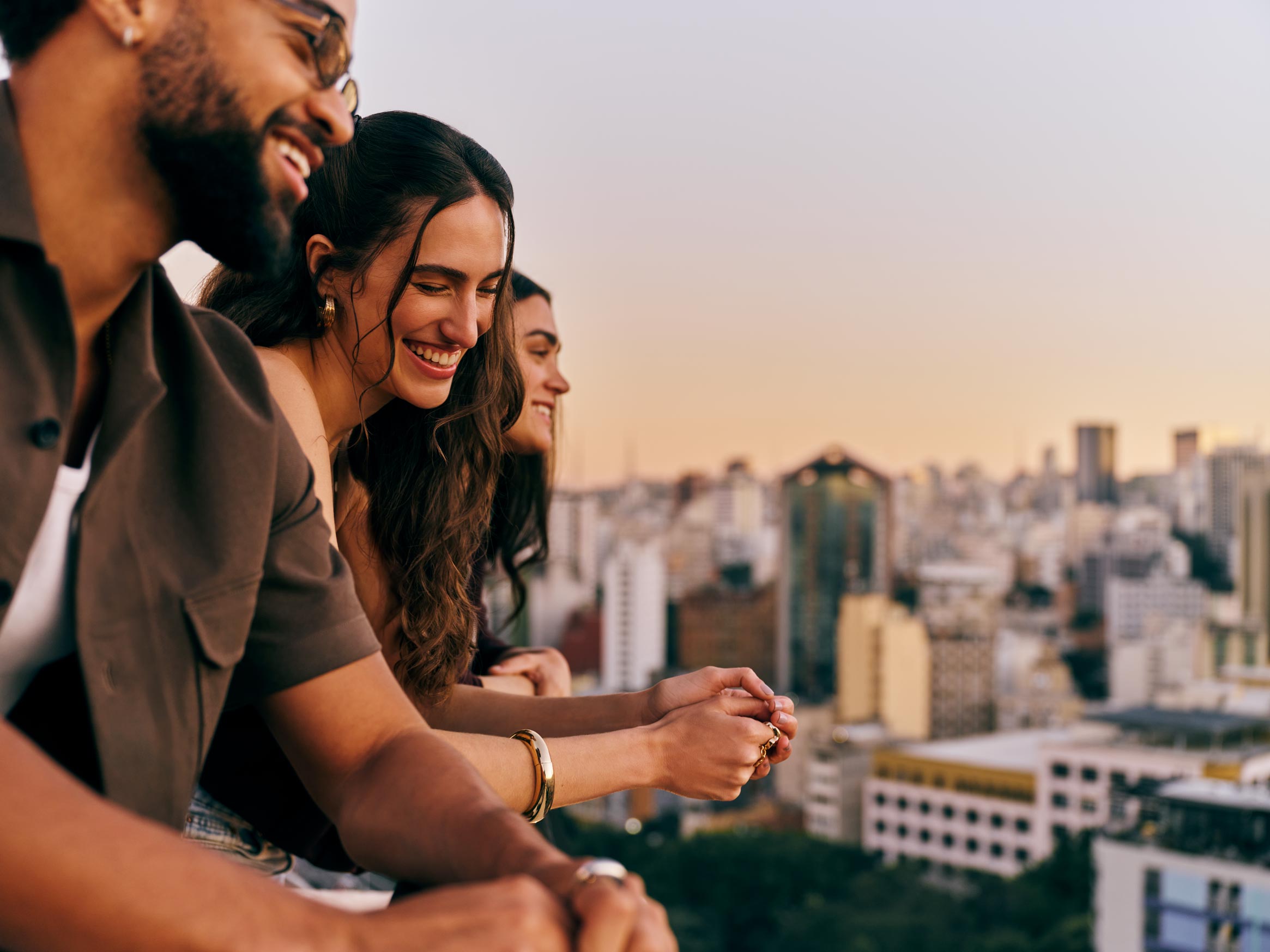 Três amigos aparecem sorrindo de perfil enquanto apreciam a vista de uma cidade a partir de um terraço durante o pôr do sol.