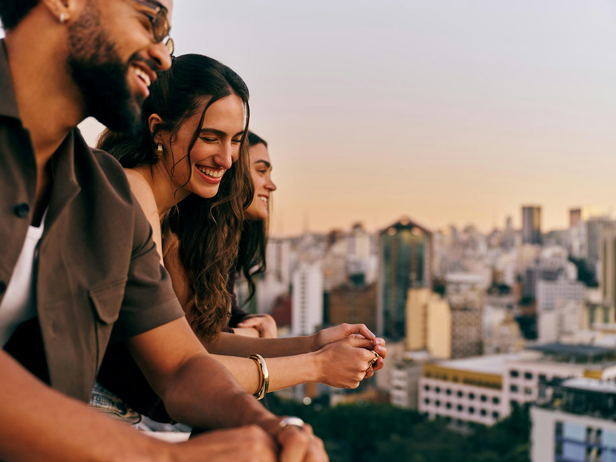 Três amigos sorrindo em um rooftop com vista da cidade.