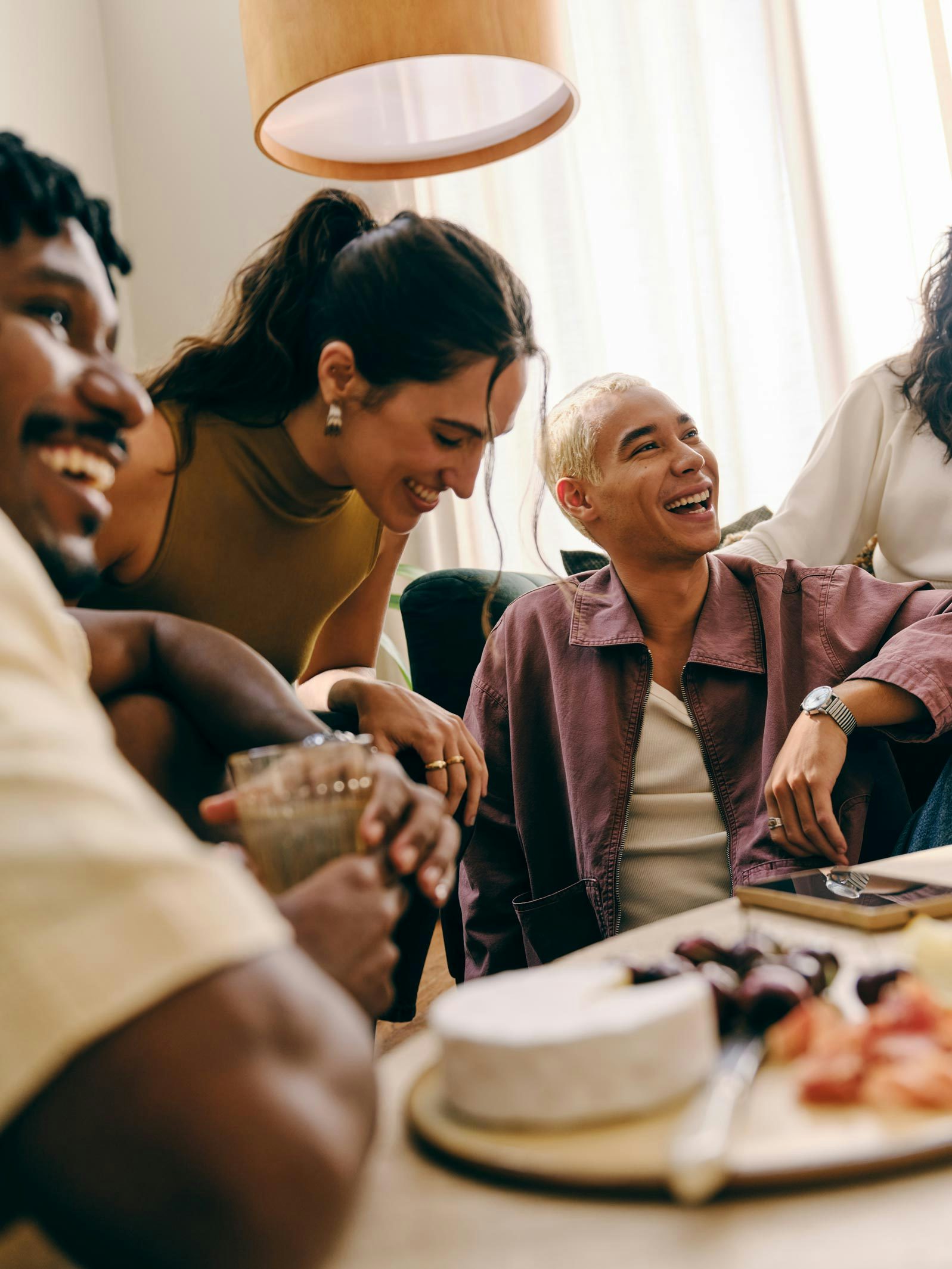 Um grupo de amigos reunido na sala de casa ao redor de uma mesa de centro com petiscos, como uma tábua de queijos e frutas.