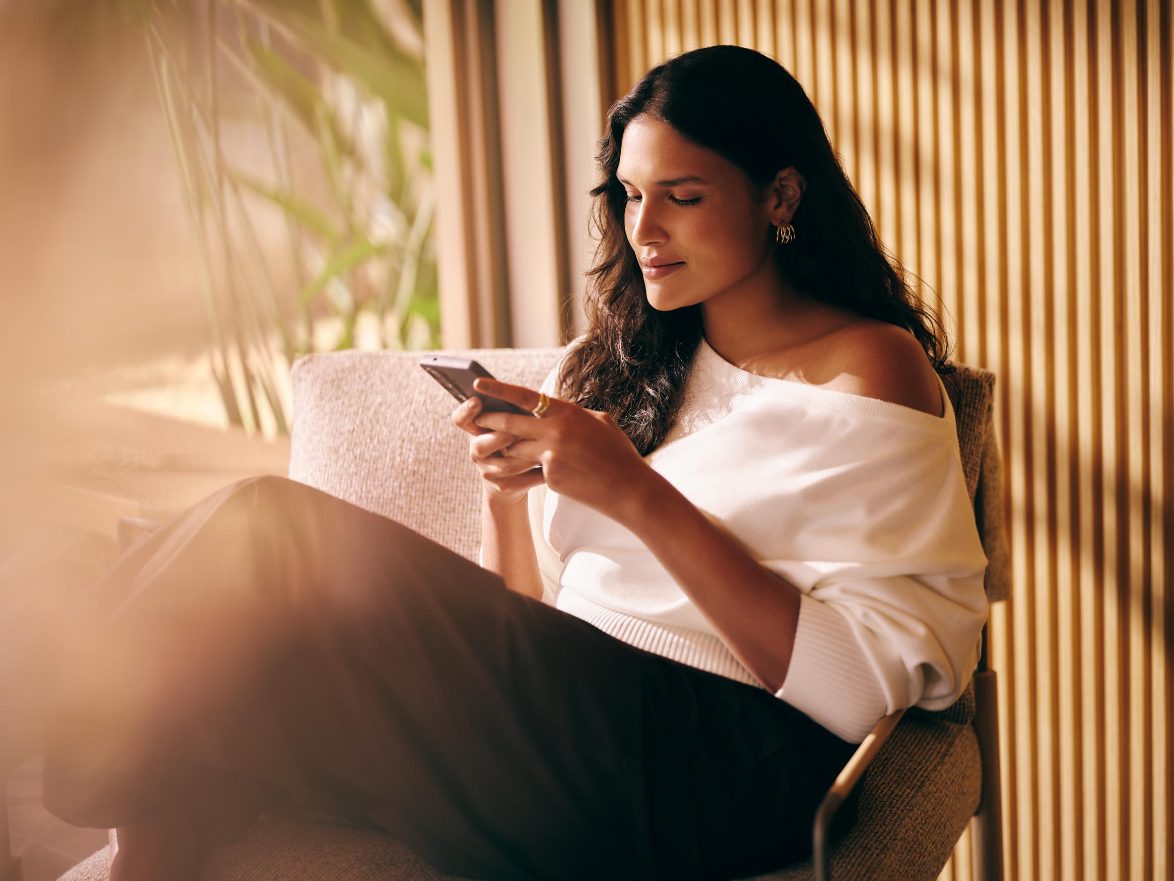 Uma mulher com blusa branca caída sobre o ombro relaxa sentada em uma poltrona enquanto mexe no celular.
