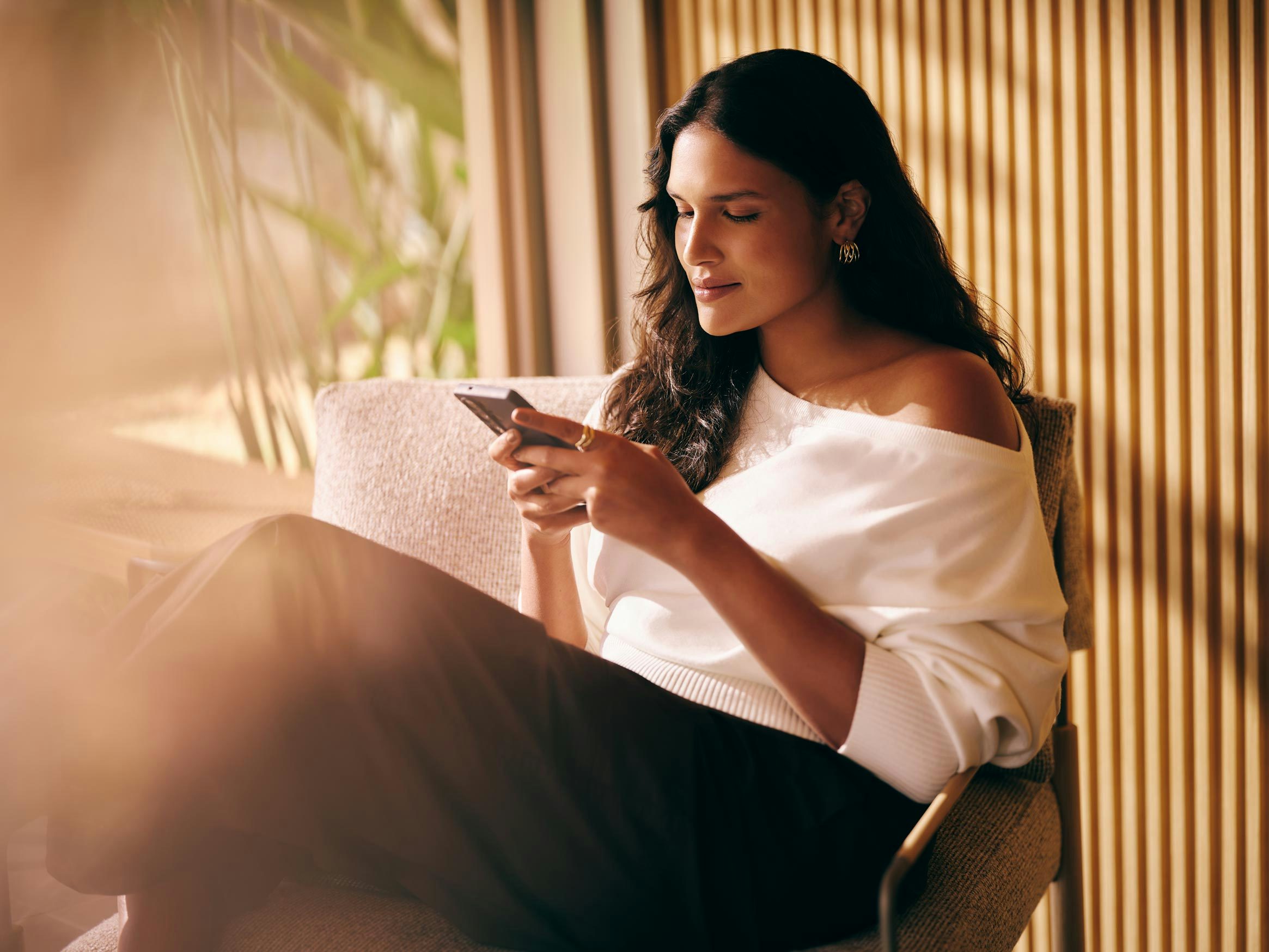 Uma mulher com blusa branca caída sobre o ombro relaxa sentada em uma poltrona enquanto mexe no celular.