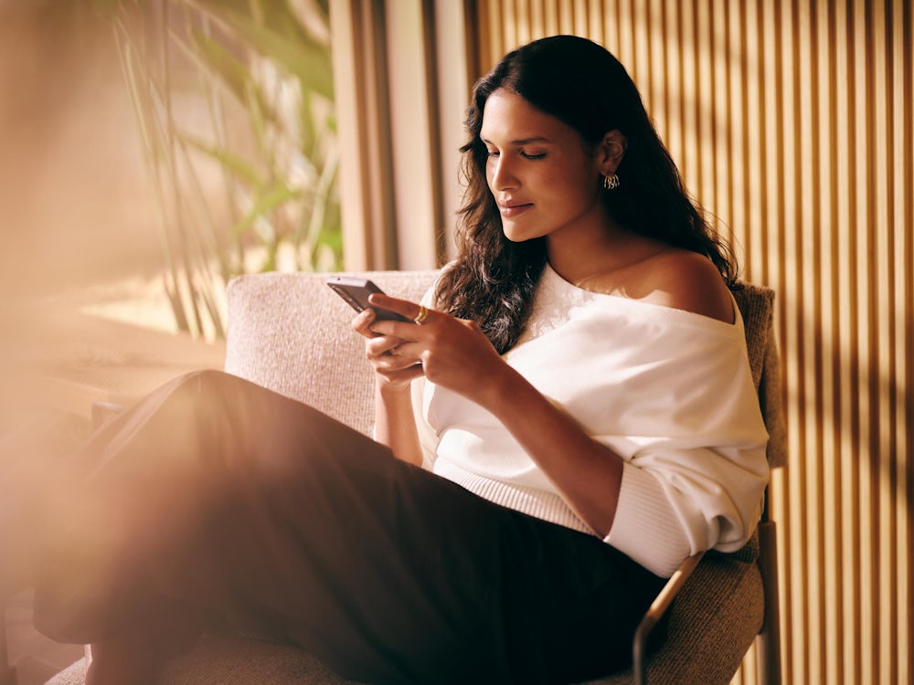 Mulher sentada em cadeira, olhando para celular com expressão tranquila.
