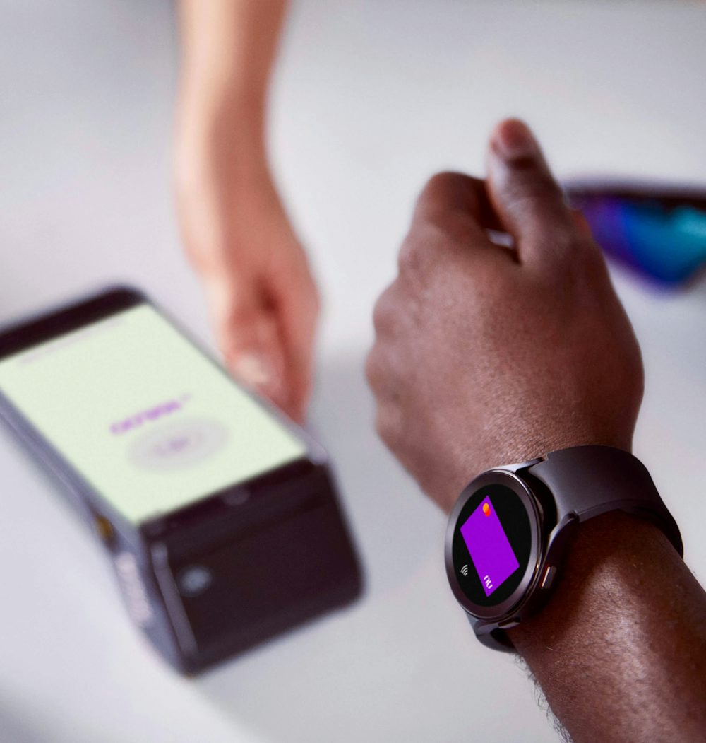 Braço de uma pessoa negra usando um smartwatch para efetuar o pagamento com seu cartão virtual Nubank por aproximação, do outro lado a mão de uma pessoa branca segurando uma máquina de cartão de crédito em cima de uma mesa com um óculos e um capacete de ciclista em cada lado.
