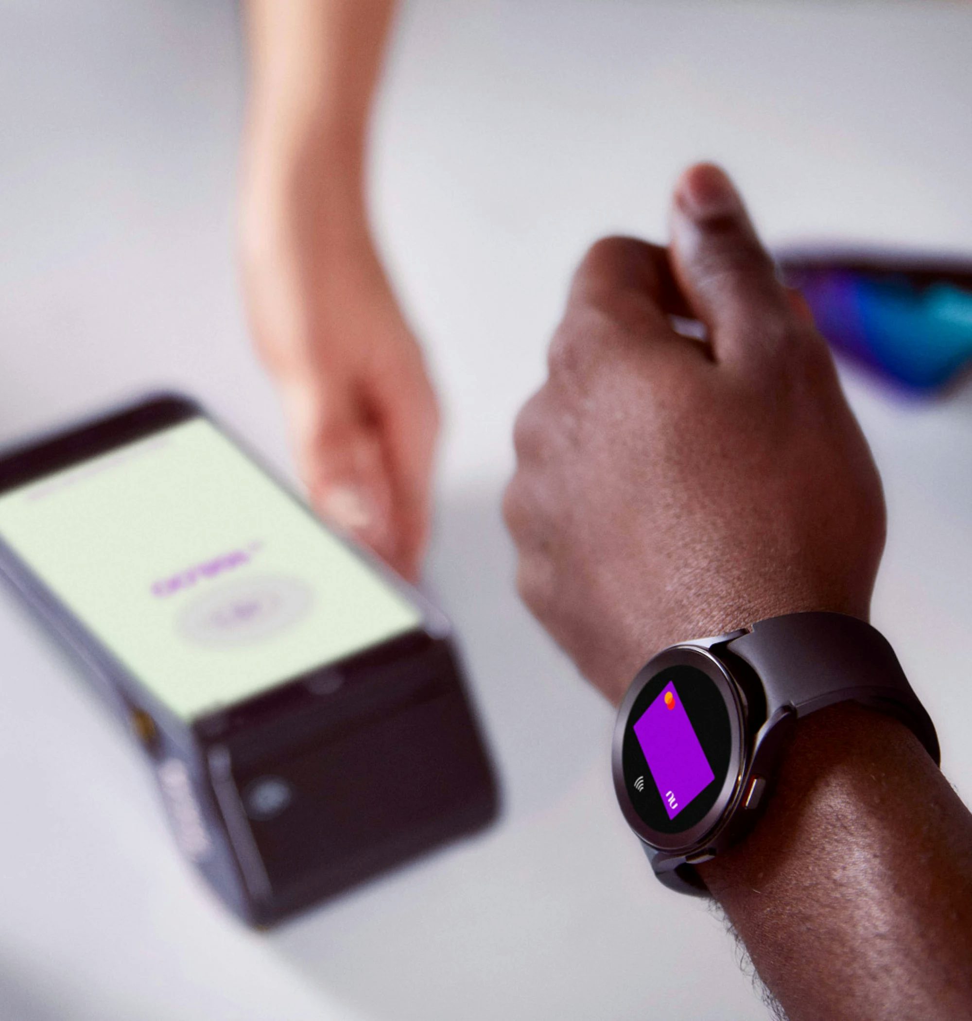 Braço de uma pessoa negra usando um smartwatch para efetuar o pagamento com seu cartão virtual Nubank por aproximação, do outro lado a mão de uma pessoa branca segurando uma máquina de cartão de crédito em cima de uma mesa com um óculos e um capacete de ciclista em cada lado.