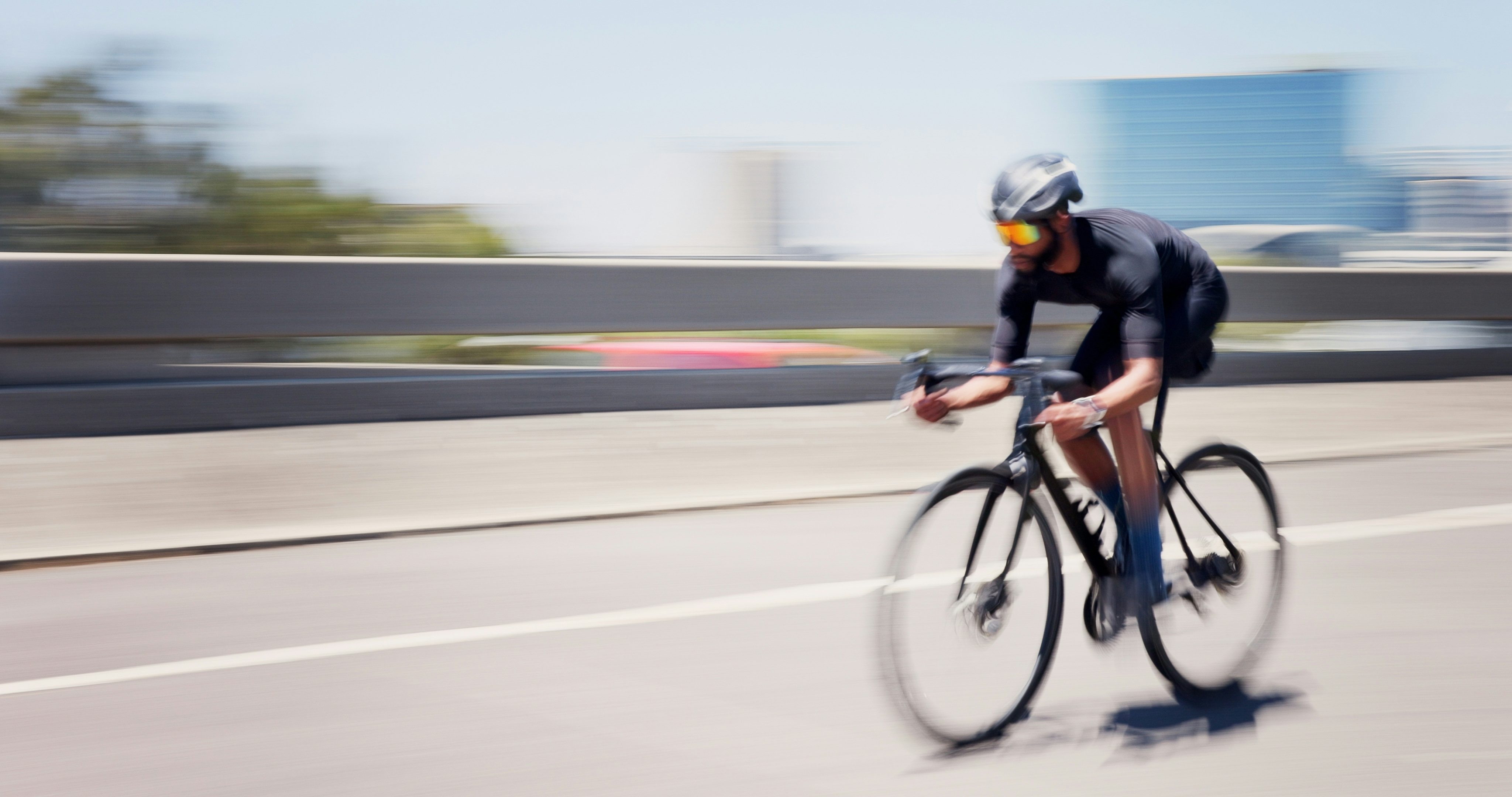 Um ciclista usando uniforme preto, capacete e óculos espelhados coloridos pedala em uma bicicleta de corrida por uma estrada urbana.