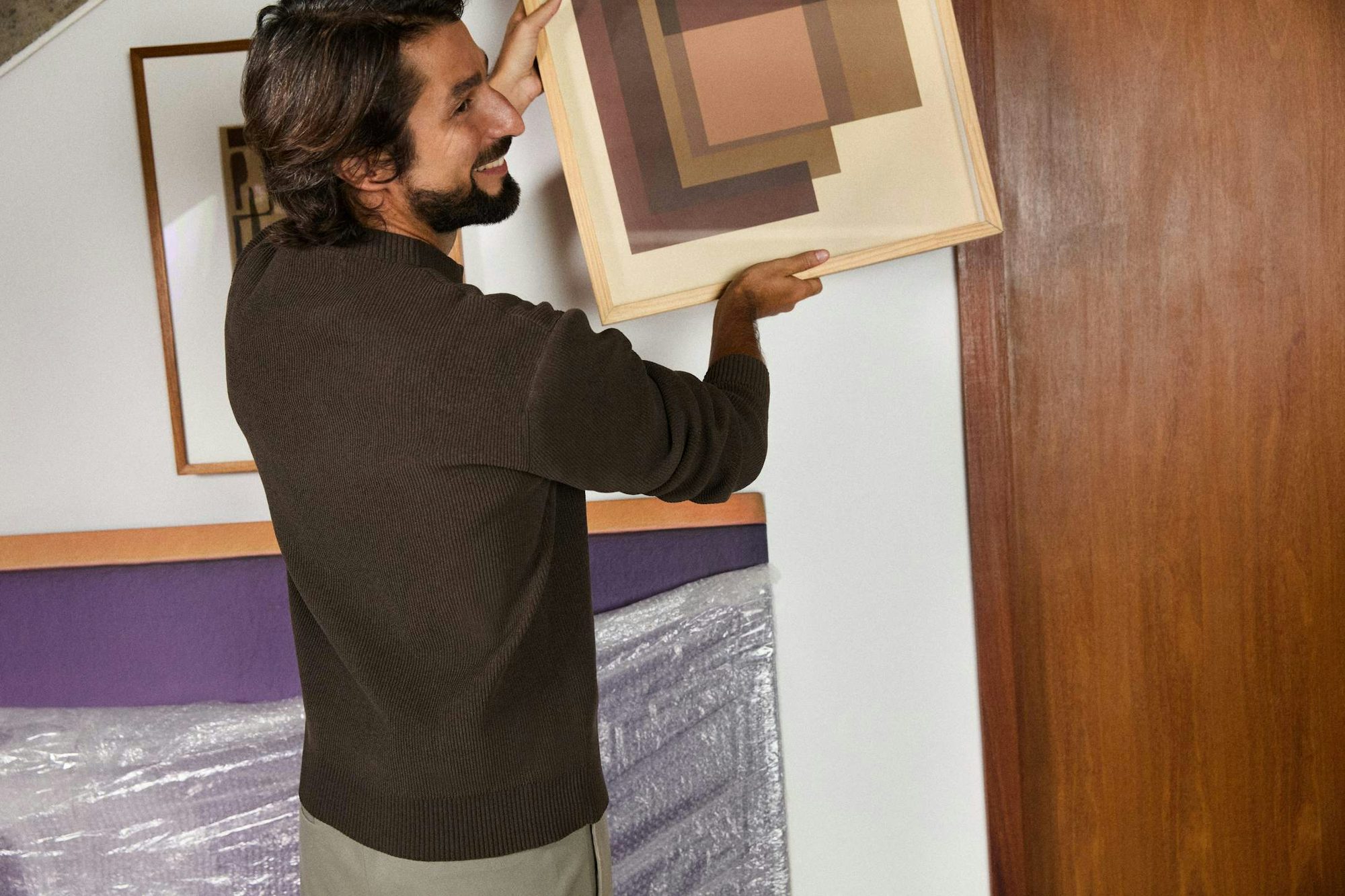 Um homem de perfil, vestindo blusa de tricô marrom, segura um quadro com arte geométrica abstrata na parede, em um momento de decoração de uma casa nova durante uma mudança.