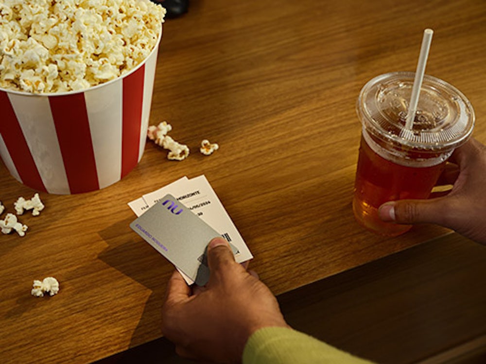 Na foto podemos ver a mão de um homem realizando o pagamento de uma pipoca e refrigerantes no cinema com o cartão virtual Nubank Croma em seu celular