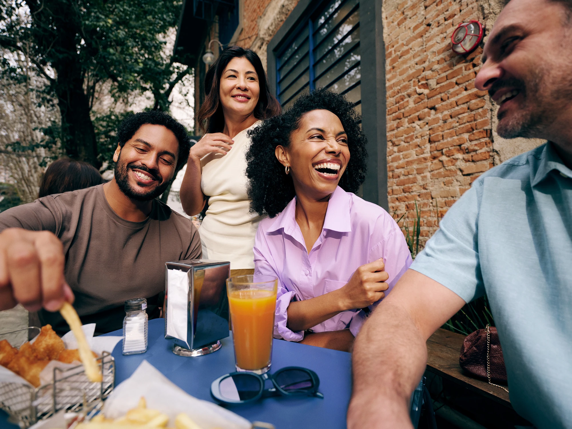 Grupo de amigos formado por 2 mulheres e dois homens sorrindo, sentados próximos a uma mesa em um ambiente que parece ser um bar ou lanchonete. Na mesa podemos ver suco, batatas fritas e guardanapos.