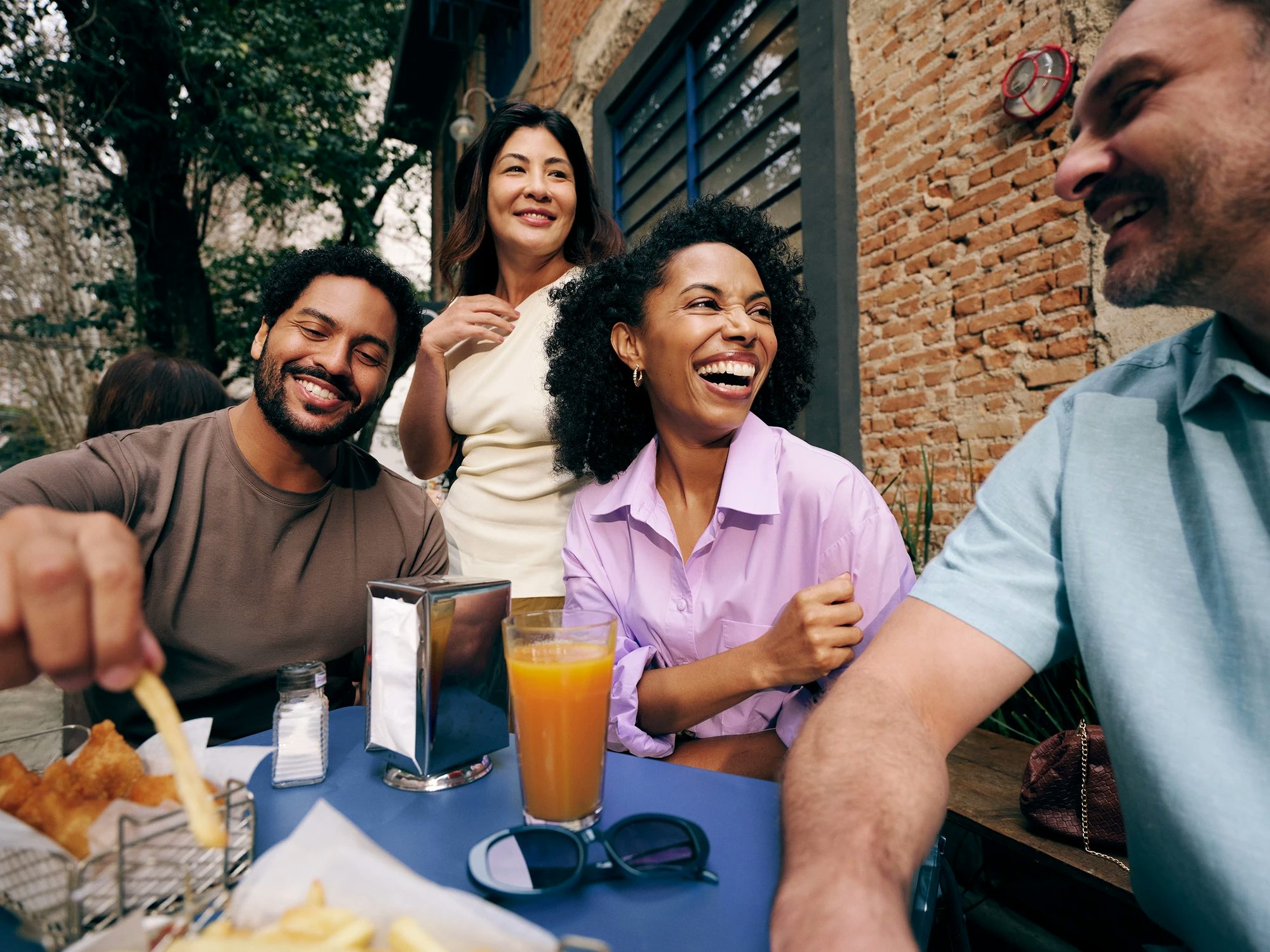 Grupo de amigos formado por 2 mulheres e dois homens sorrindo, sentados próximos a uma mesa em um ambiente que parece ser um bar ou lanchonete. Na mesa podemos ver suco, batatas fritas e guardanapos.