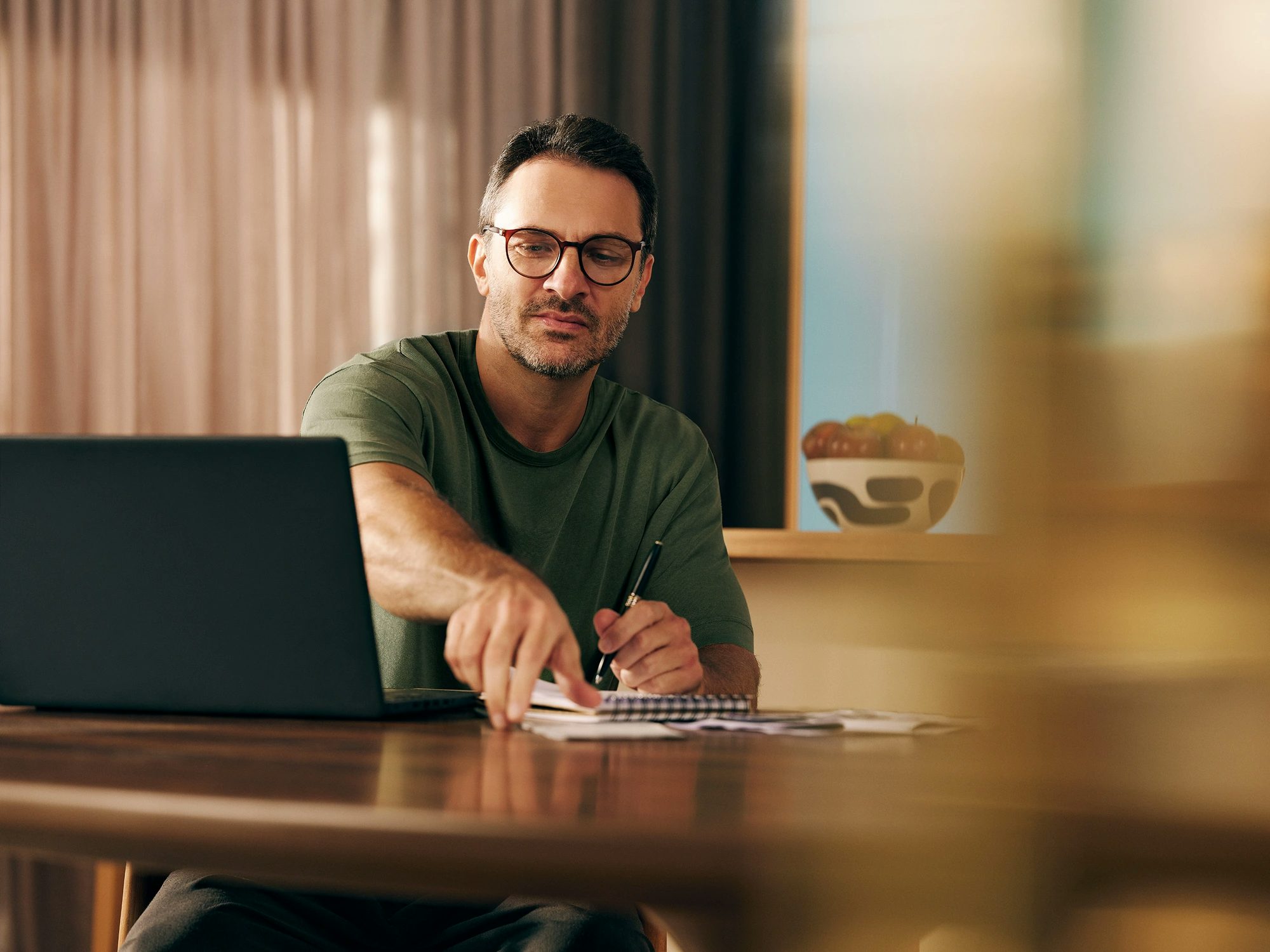 Homem sentado próximo a uma mesa, olhando para um notebook, enquanto faz anotações em um caderno