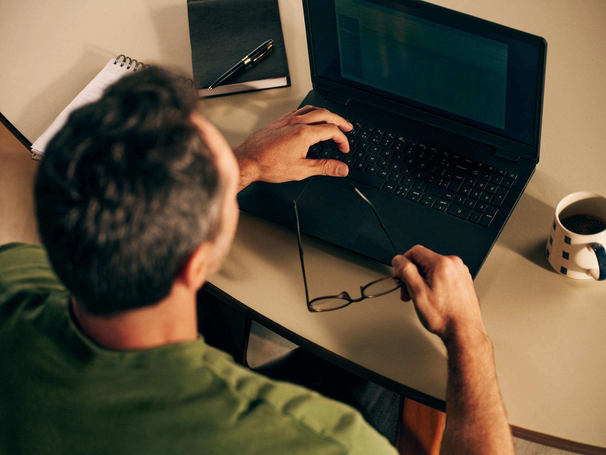 Homem sentado próximo a uma mesa, olhando para um notebook, enquanto faz anotações em um caderno
