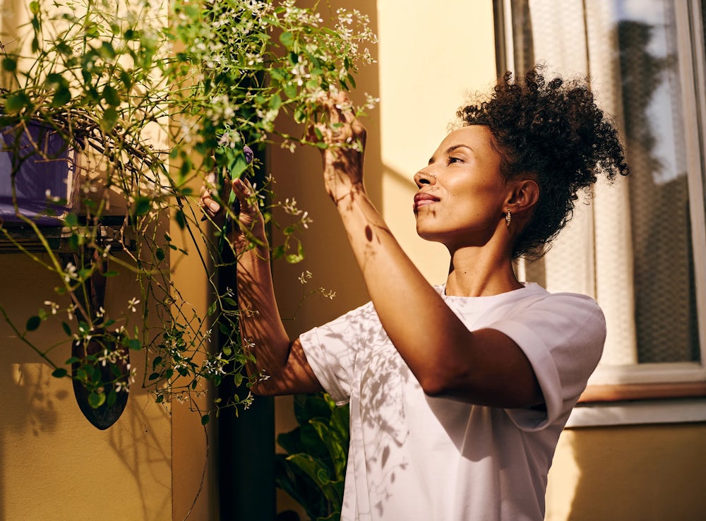 Mulher negra com leve sorriso, em pé em seu quintal, cuidando de uma planta