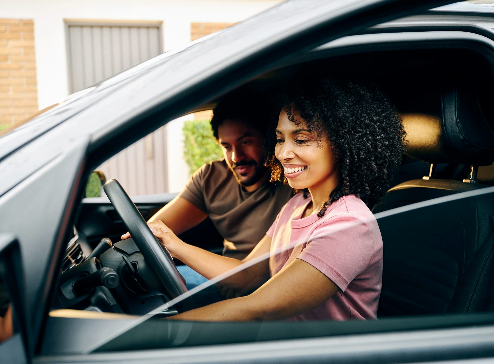 Casal dentro de um carro. A mulher está no banco do motorista, abrindo a porta do carro, enquanto o homem está no banco do passageiro. Ambos sorriem.