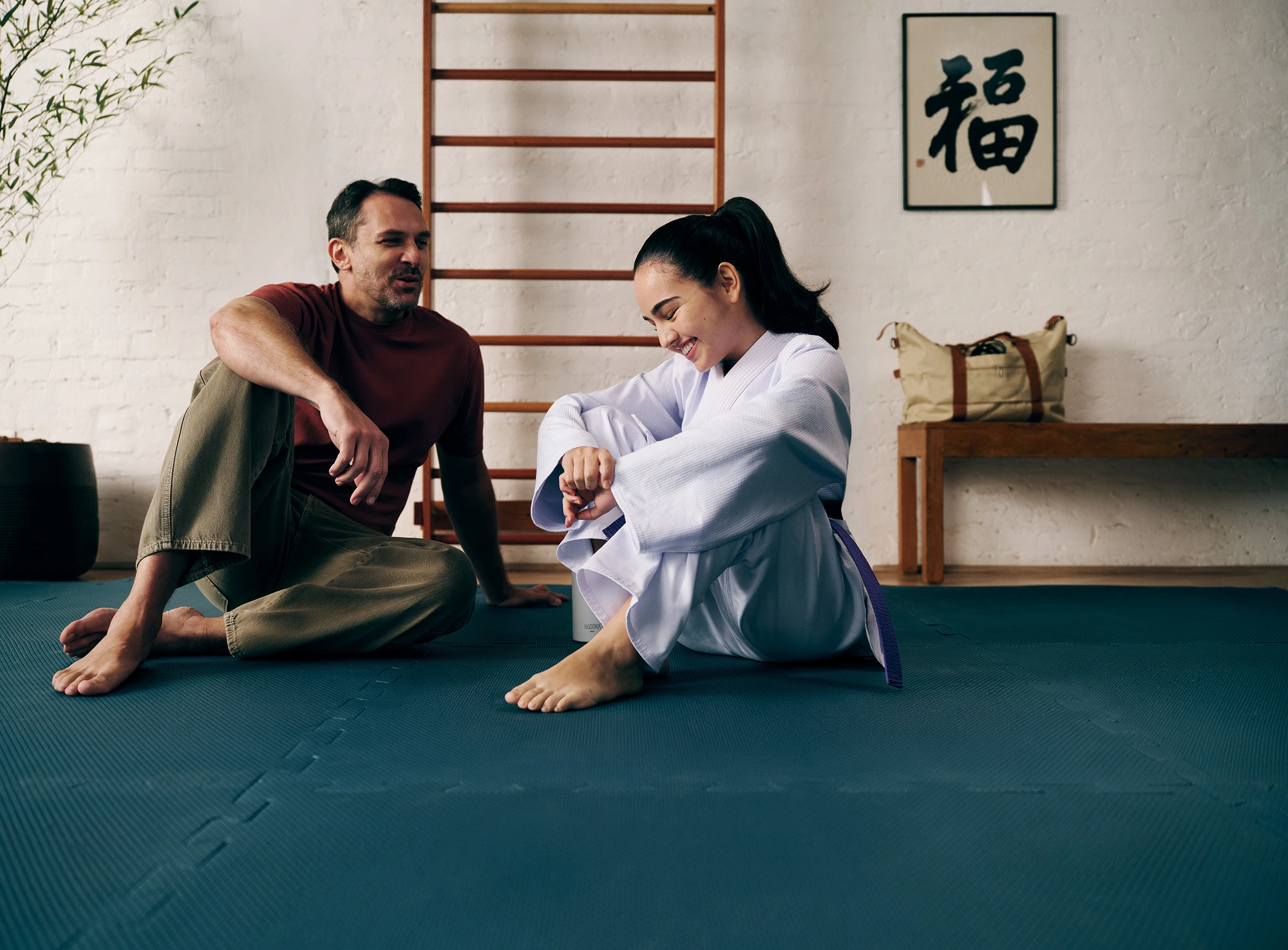 Pai e filha estão sentados em um tatame de Judo. A garota, jovem, está com trajes dessa arte marcial e seu pai, um homem de 40 anos, esta com calça jeans e camiseta.