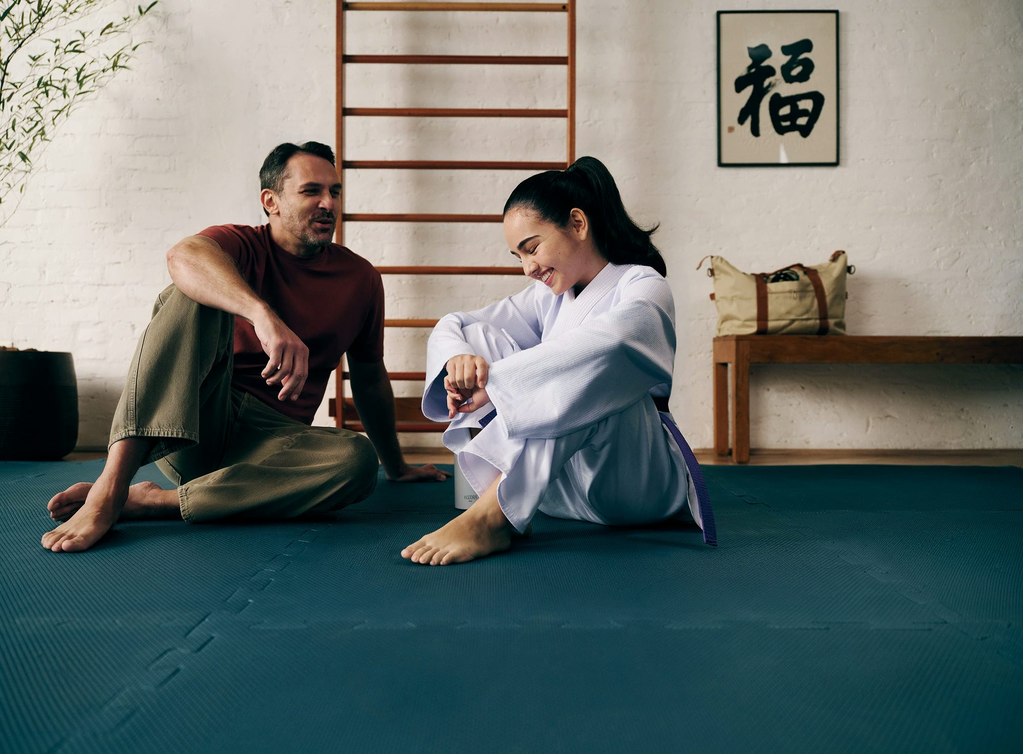 Pai e filha estão sentados em um tatame de Judo. A garota, jovem, está com trajes dessa arte marcial e seu pai, um homem de 40 anos, esta com calça jeans e camiseta.