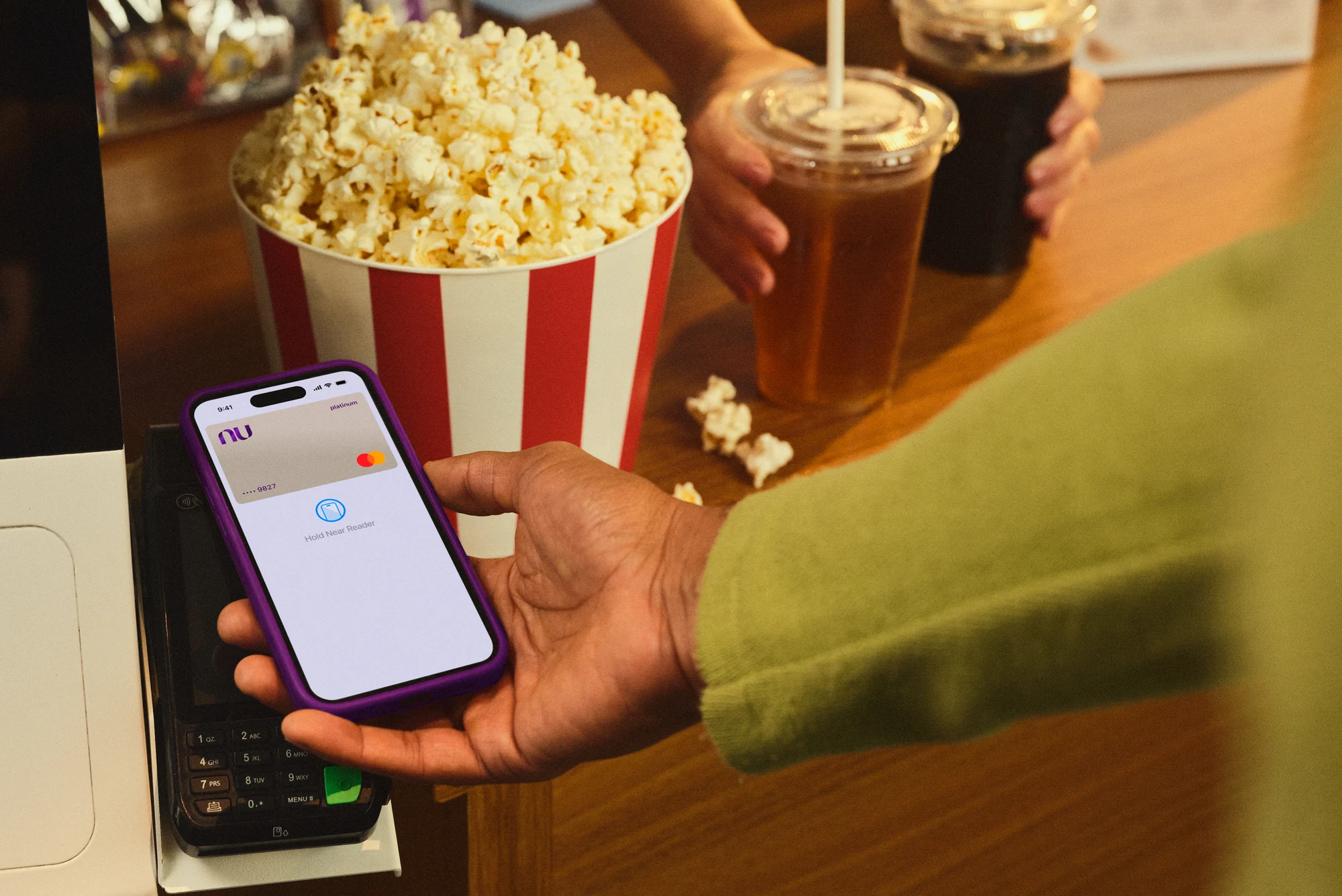 Na foto podemos ver a mão de um homem realizando o pagamento de uma pipoca e refrigerantes no cinema com o cartão virtual Nubank Croma em seu celular