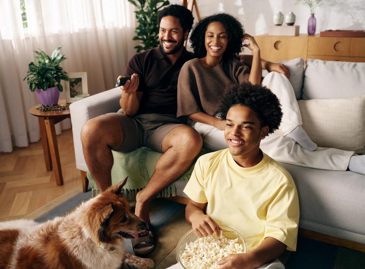 Família reunida com pai e mãe sentados ao sofá, filho sentado ao chão com balde de pipoca no colo e cachorro próximo a ele.