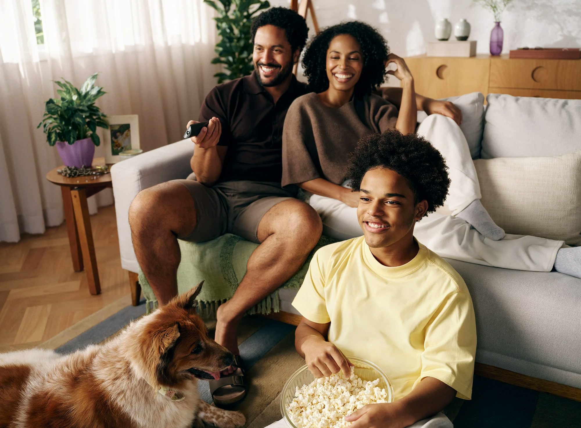 Família reunida com pai e mãe sentados ao sofá, filho sentado ao chão com balde de pipoca no colo e cachorro próximo a ele.