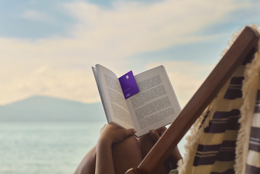 Uma mulher deitada em uma cadeira de praia segurando um livro aberto com o cartão Nubank Ultravioleta no meio