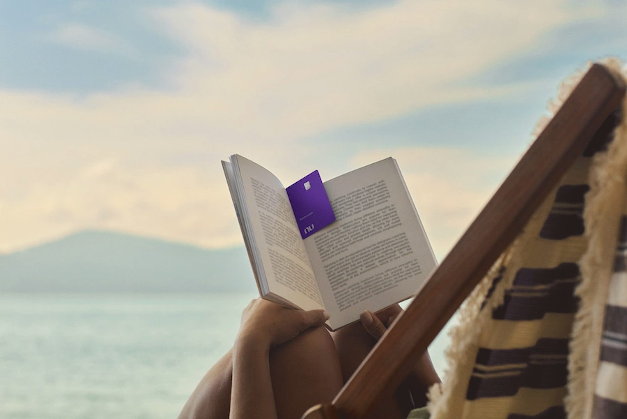 Uma mulher deitada em uma cadeira de praia segurando um livro aberto com o cartão Nubank Ultravioleta no meio