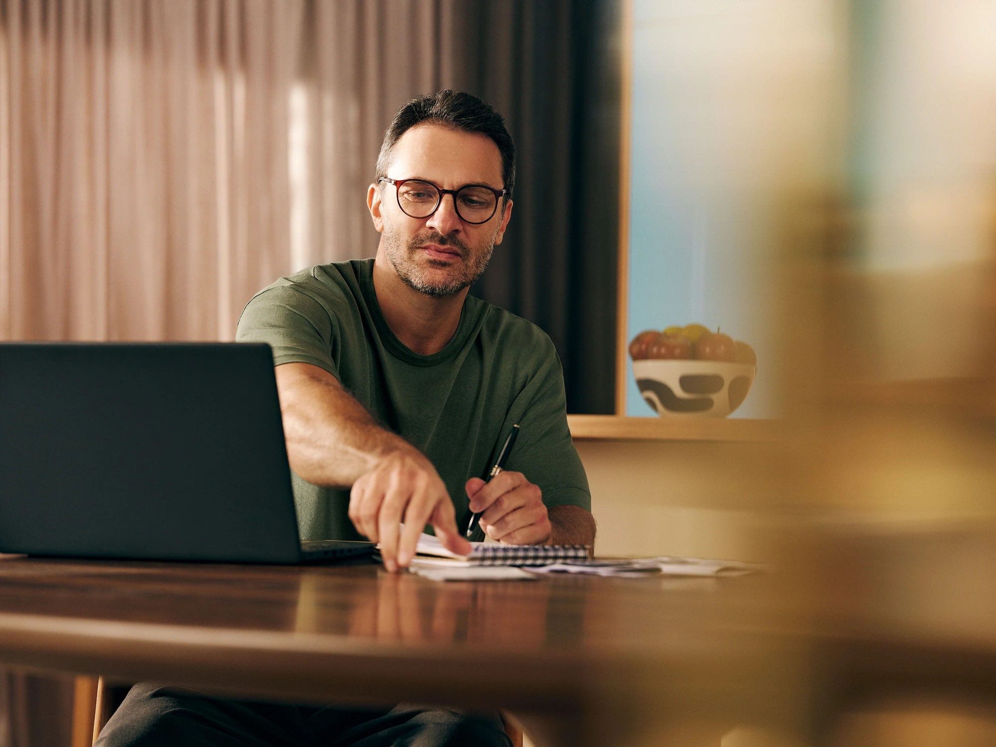 Homem sentado próximo a uma mesa, olhando para um notebook, enquanto faz anotações em um caderno
