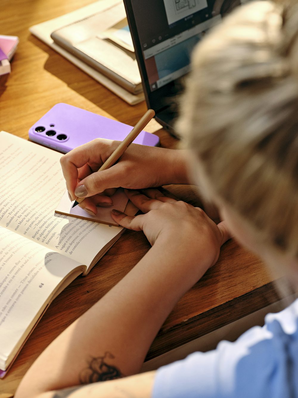Uma pessoa aparece estudando em uma mesa de madeira, fazendo anotações com lápis em um pequeno bloco sobre um livro aberto. Ao redor, há um celular com capinha lilás e um notebook aberto, sob uma iluminação quente e acolhedora.