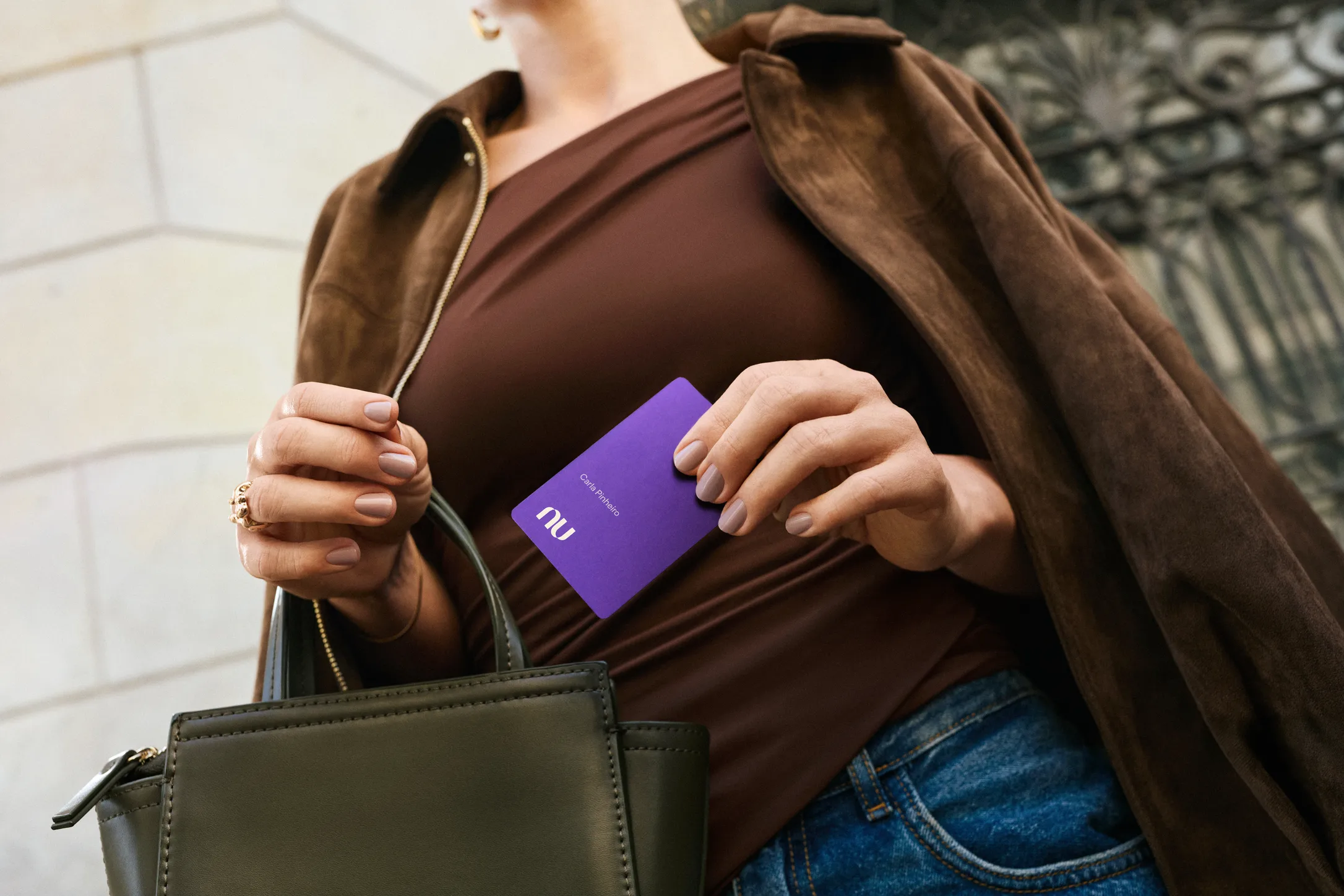 Detalhe focado nas mãos de uma mulher segurando o cartão Nubank Ultravioleta. Ela veste um casaco marrom de camurça, blusa escura e calça jeans, segurando também uma bolsa de mão verde-oliva.