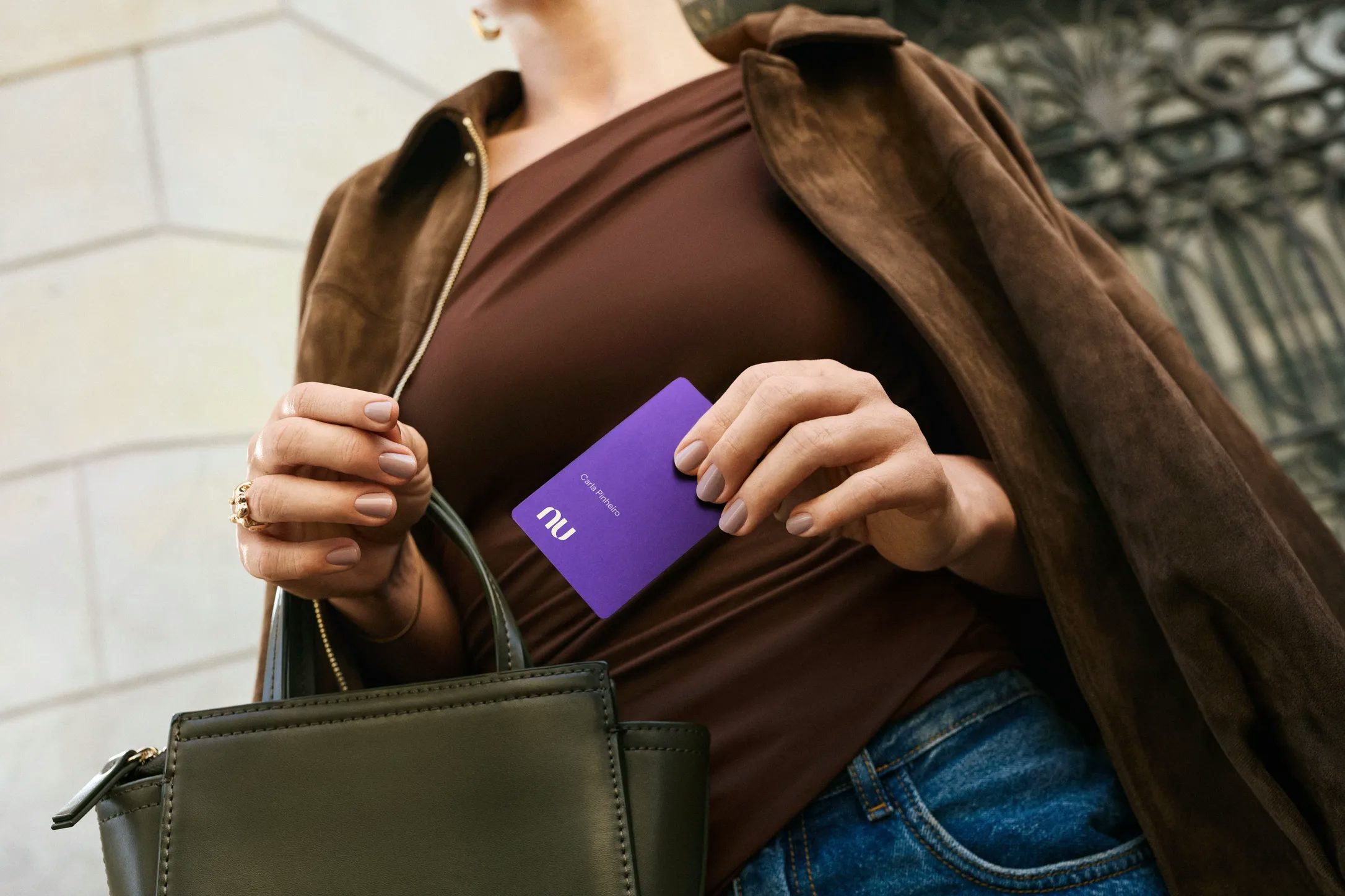 Detalhe focado nas mãos de uma mulher segurando o cartão Nubank Ultravioleta. Ela veste um casaco marrom de camurça, blusa escura e calça jeans, segurando também uma bolsa de mão verde-oliva.