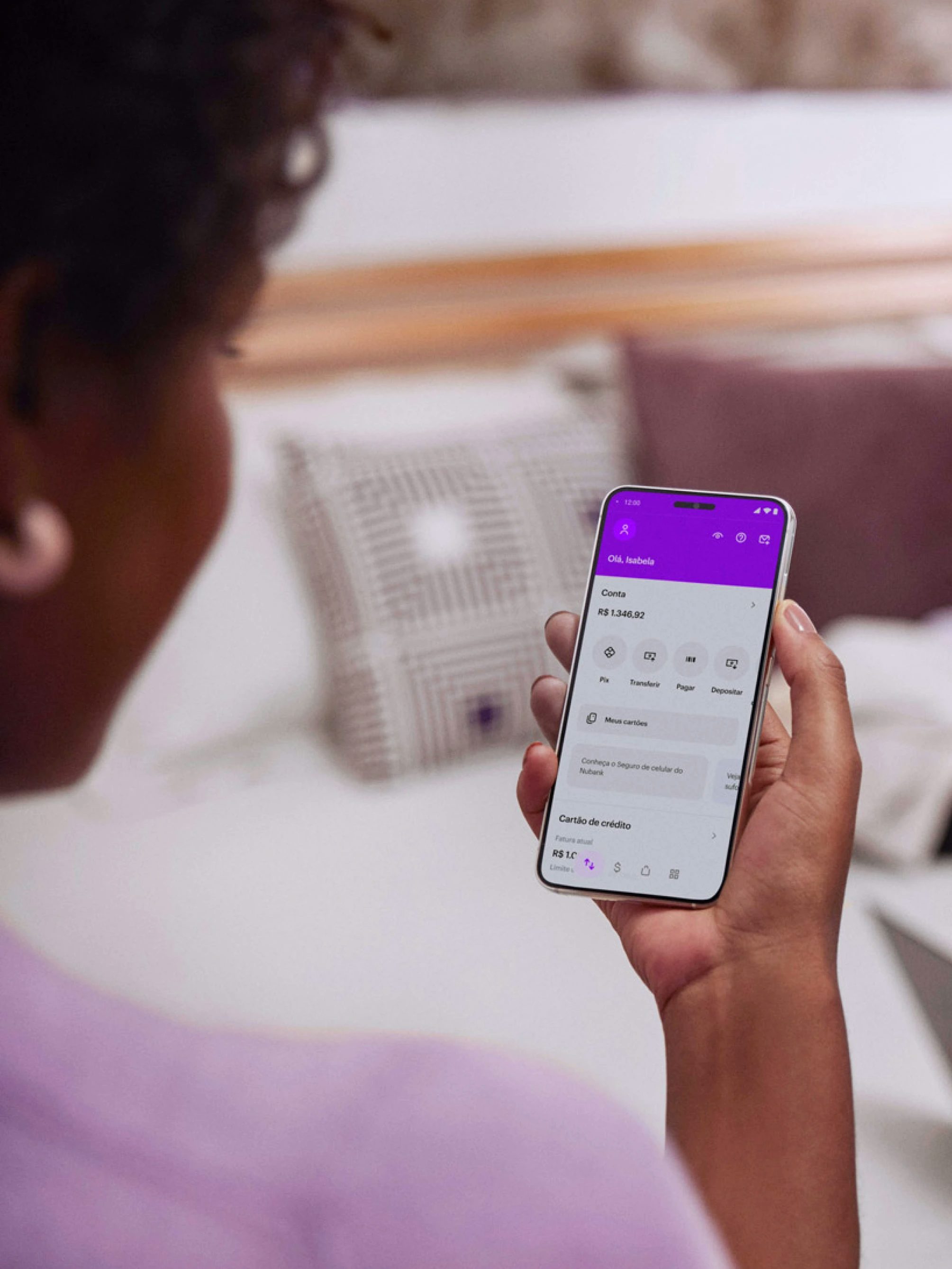 Vista das costas de uma mulher negra sentada com um celular na mão direita e visualizando sua conta no aplicativo do Nubank.
