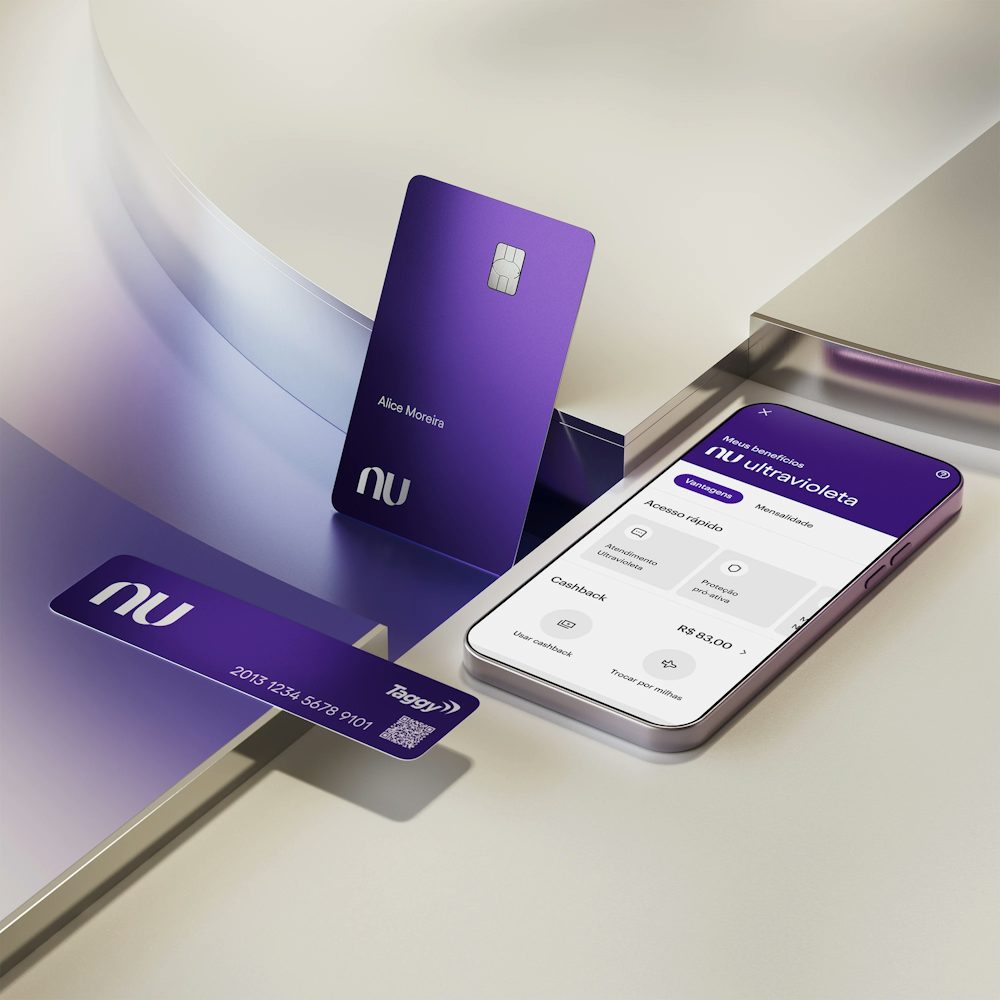 Celular com aplicativo Nubank aberto na tela ao lado de um cartão de uma tag do NuTag. No canto superior, a extremidade de um cartão ultravioleta. Os elementos estão apoiados em uma forma geométrica.
