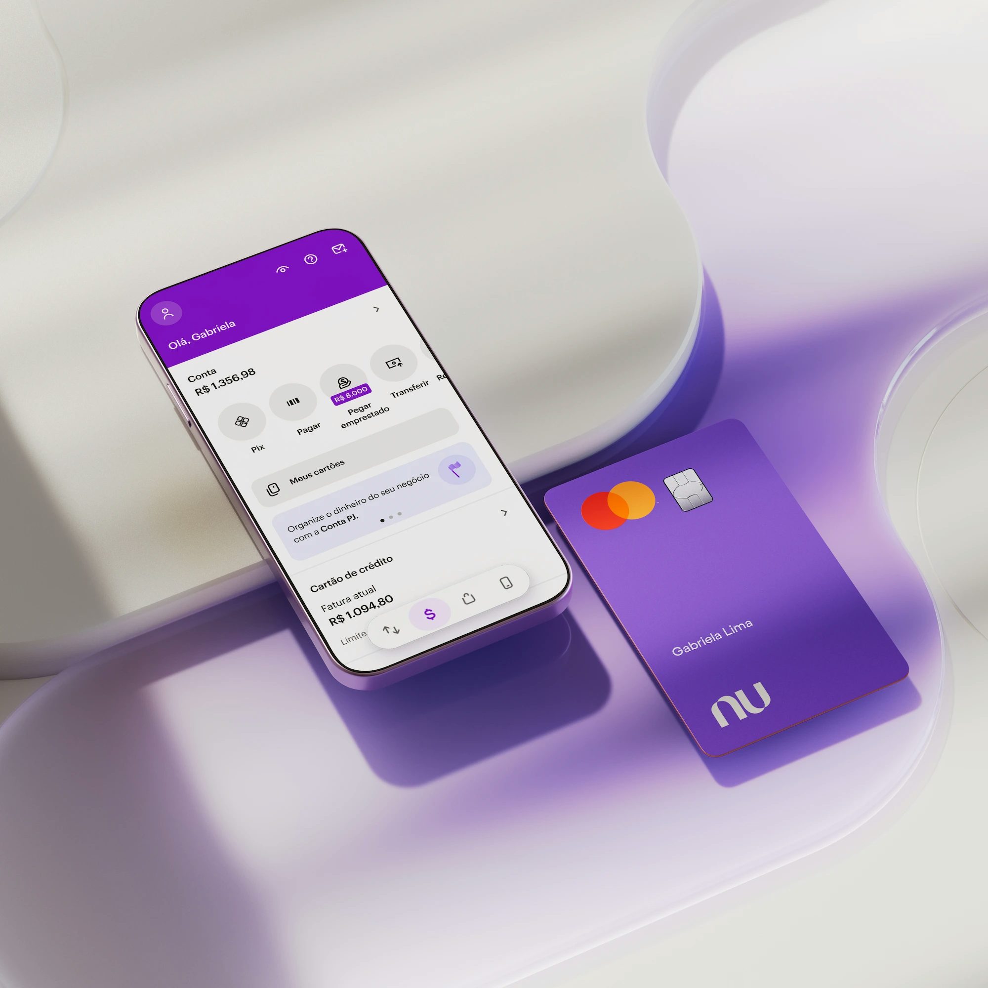 Celular com aplicativo Nubank aberto na área inicial com o saldo da conta na parte superior da tela. Na parte inferior do aplicativo na tela do celular, informações sobre Cartão de crédito. O celular e o cartão estão deitados apoiados em uma forma geométrica branca.