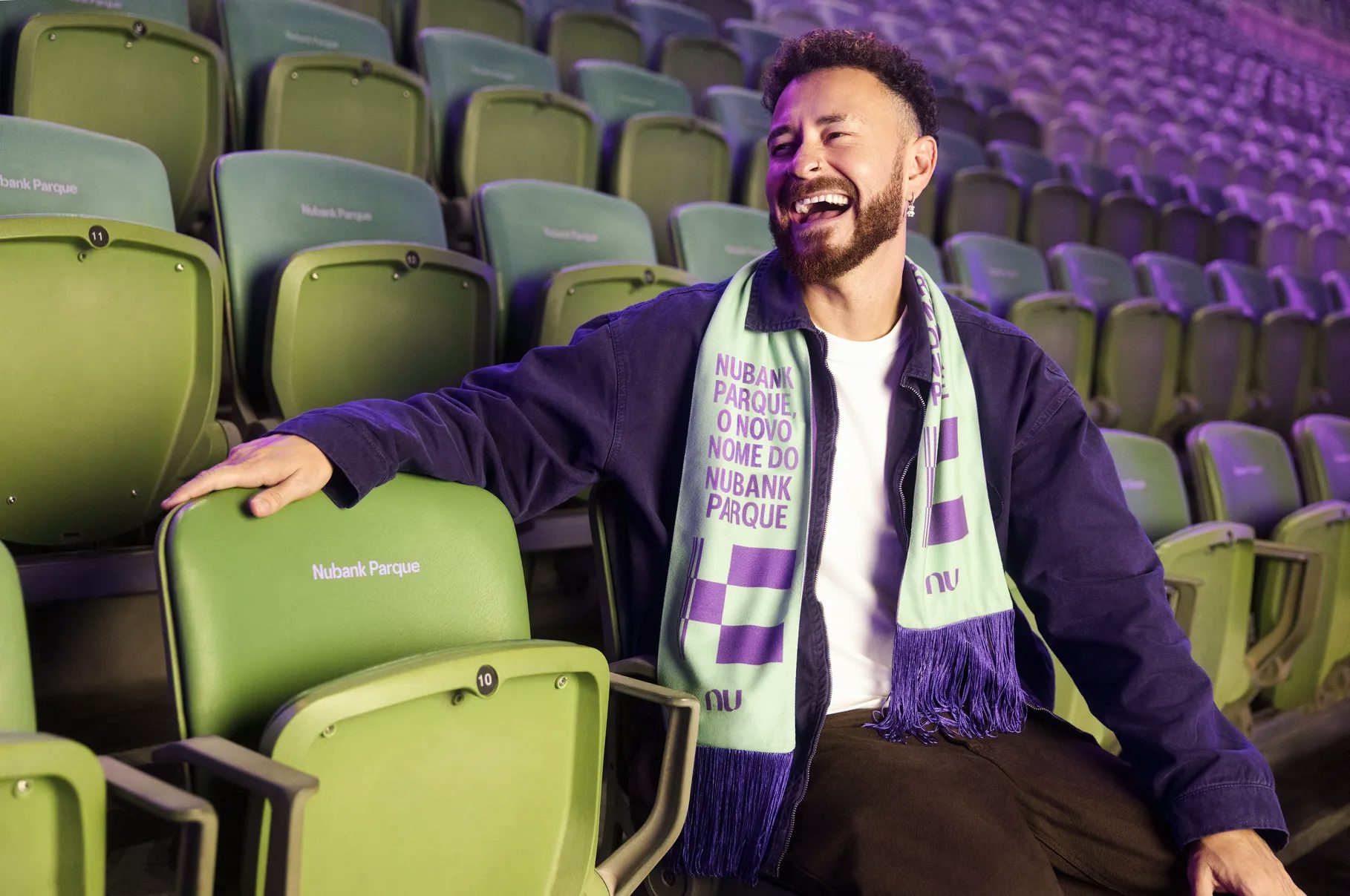 Fred sorrindo sentado na arquibancada verde. Ele usa do cachecol com o novo nome do Nubank Parque